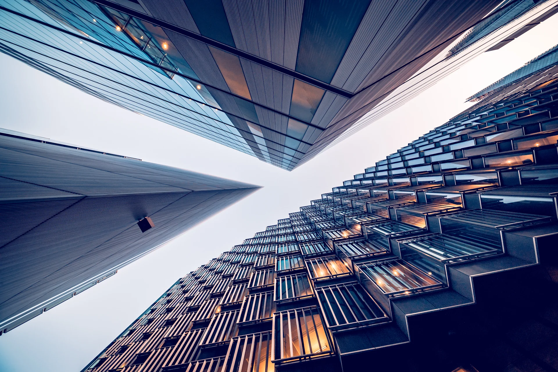 A low-angle view of two modern skyscrapers converging towards the sky, showcasing sleek glass and metal facades. The building on the left features a smooth surface with a hint of reflection, while the building on the right displays a textured pattern with illuminated windows.