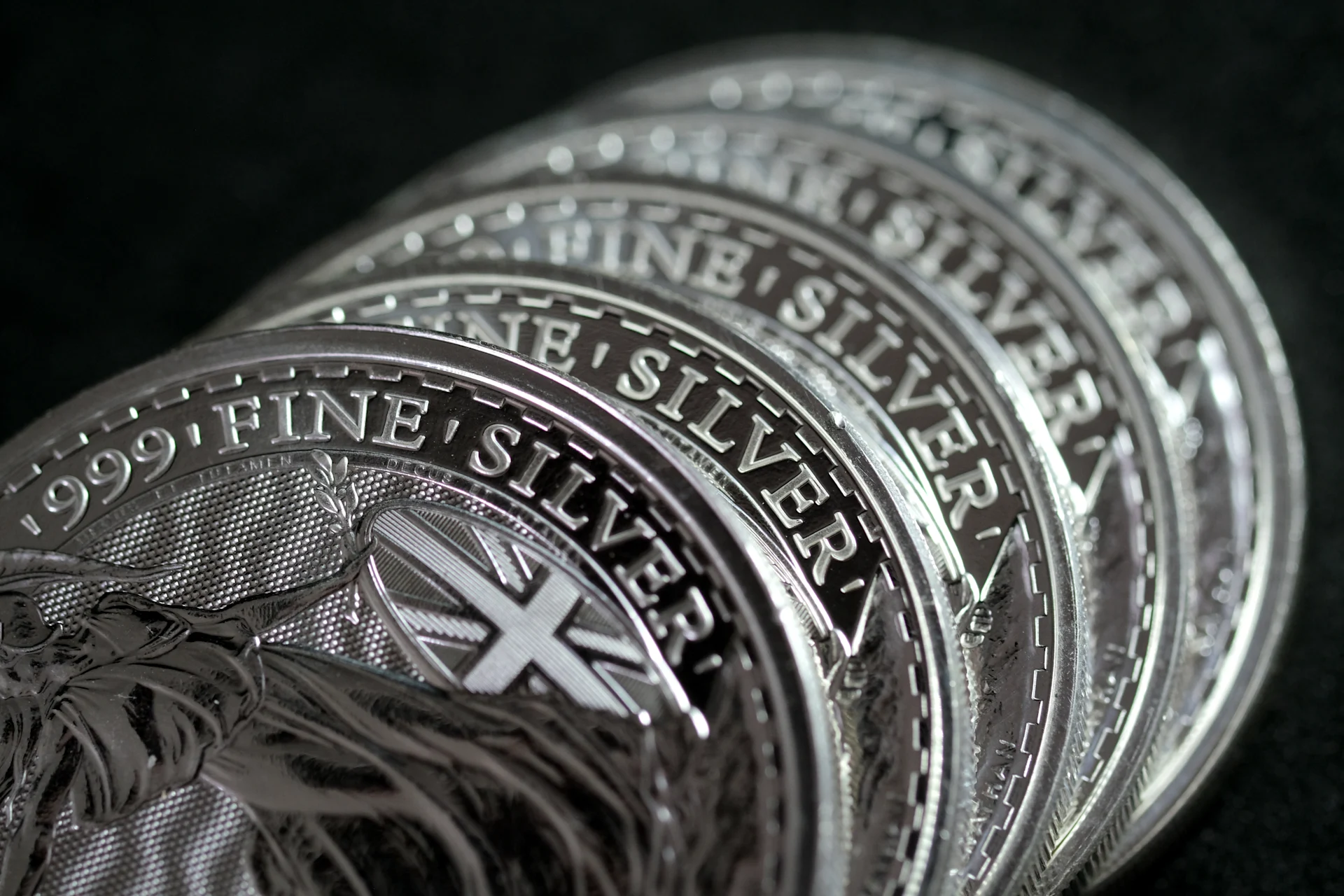 A close-up view of five silver coins arranged on a smooth black surface, reflecting light and showcasing their intricate designs.