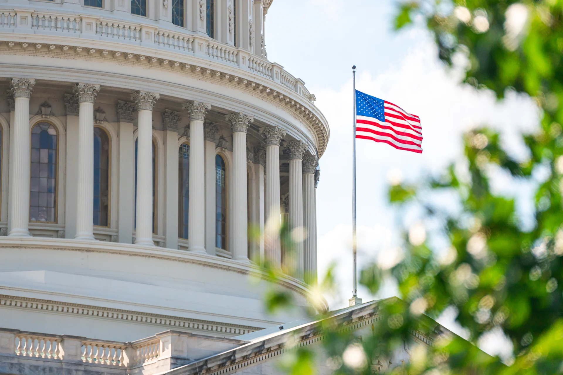 US flag at full mast with the Capitol Building prominently displayed in the background against a clear blue sky.