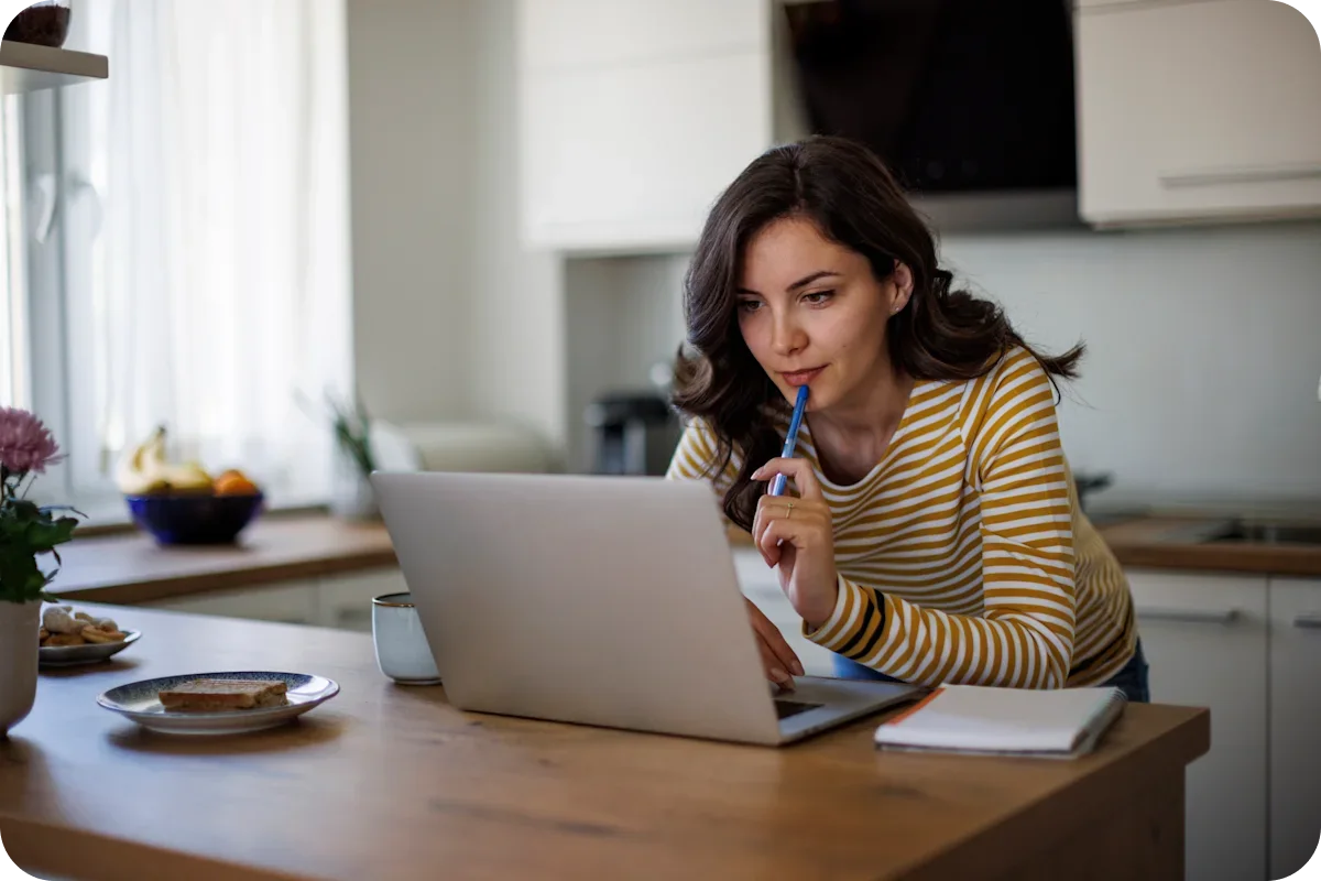 A woman seated at a table, focused on her laptop, with a notepad and pen beside her.