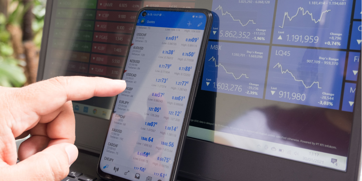 A man analysing stock market data on his MT4 account through his phone, sitting at a desk with financial documents nearby.