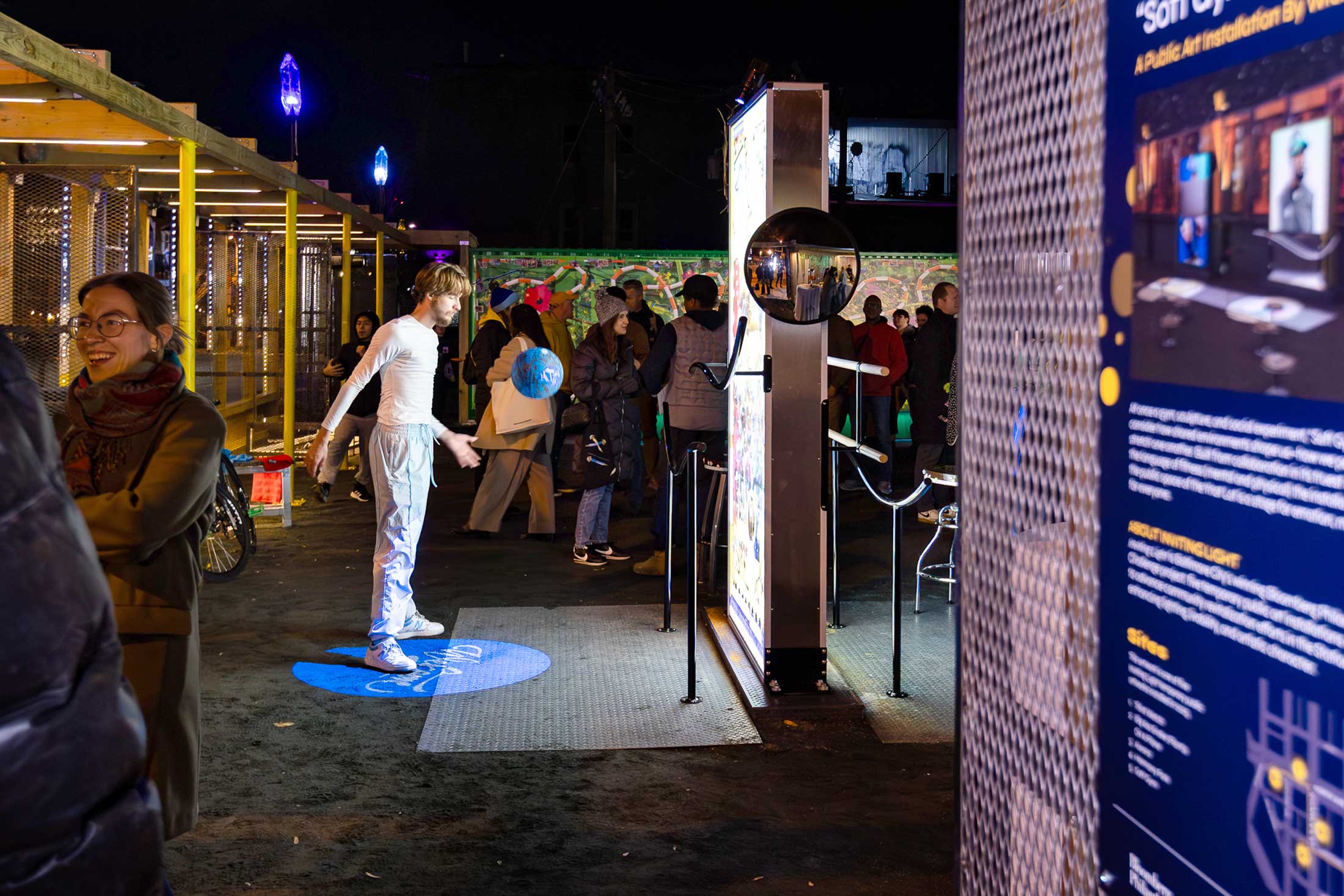 Soft Gym by Wickerham and Lomax, an Inviting Light installation at YNOT LOT in Station North Arts District. A person uses the exercise equipment in the surreal arena, surrounded by people celebrating the unveiling.