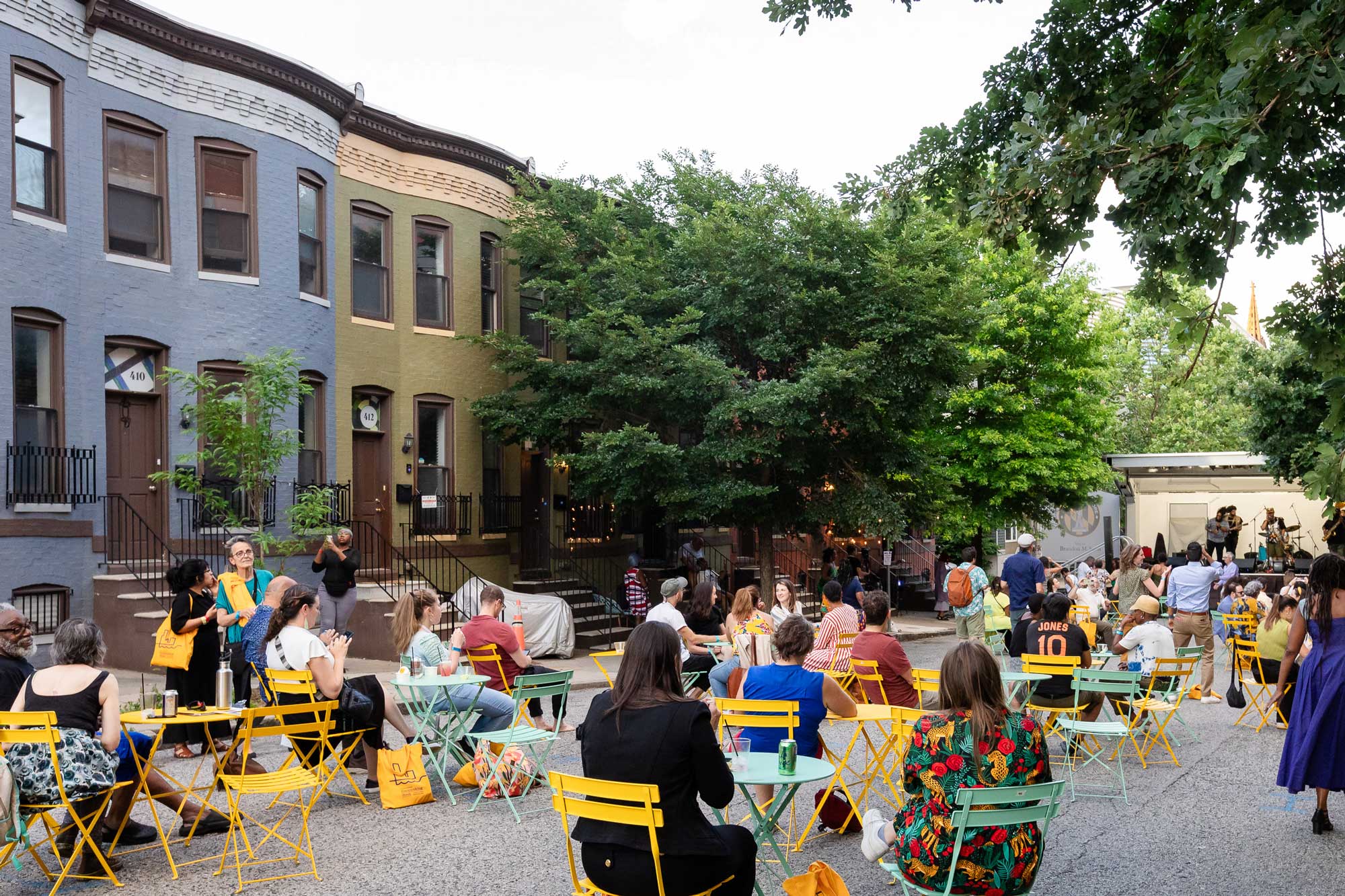 Community block party in Greenmount West, Baltimore. People sit at tables and dance to live music against a thriving backdrop of tree-lined rowhouses.