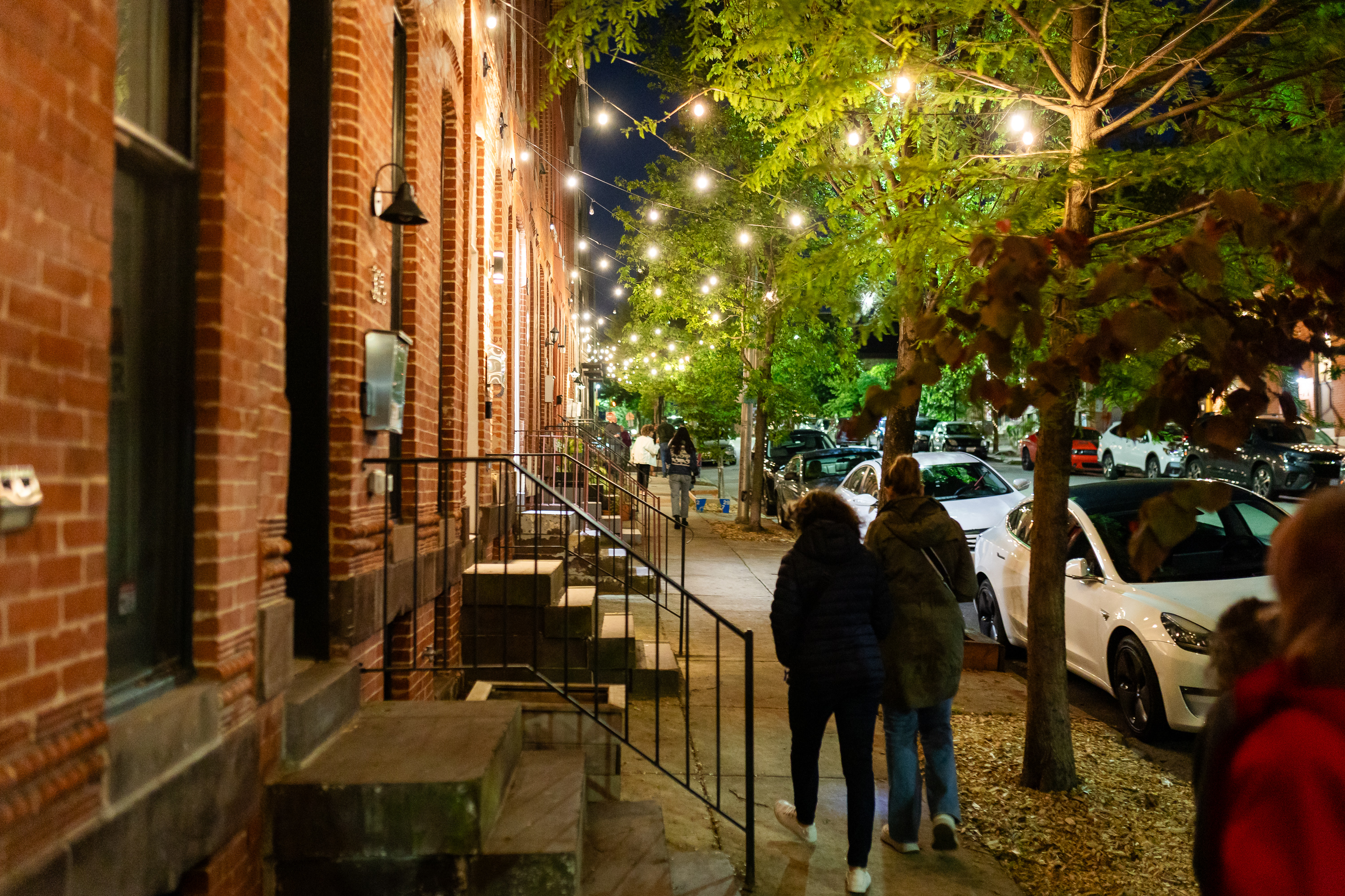 Bistro lights in trees give soft glow over sidewalk in Station North and walkers pass.
