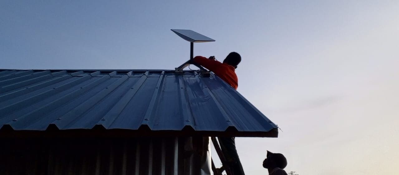 Two people work on installing a satellite dish on a corrugated metal roof at dusk. One person is perched on the rooftop, adjusting the dish, while the other stands below, looking up to provide assistance. The sky is clear with a gradual transition from light to dark.