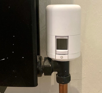 A smart radiator valve attached to a radiator pipe. The valve has a small screen and a button. The top of the cap twists to control the temperature. The button is used for pairing with the smart phone app. The screen is blank.