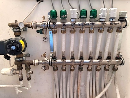 Manifold for underfloor heating system. There are 2 rows of metal rails. One has plastic caps on top. The other has gauges or meters on top. Plastic tubes come out of the bottom. Next to the rows of tubes and caps, there are several valves and dials to control the water coming in and out of the system.