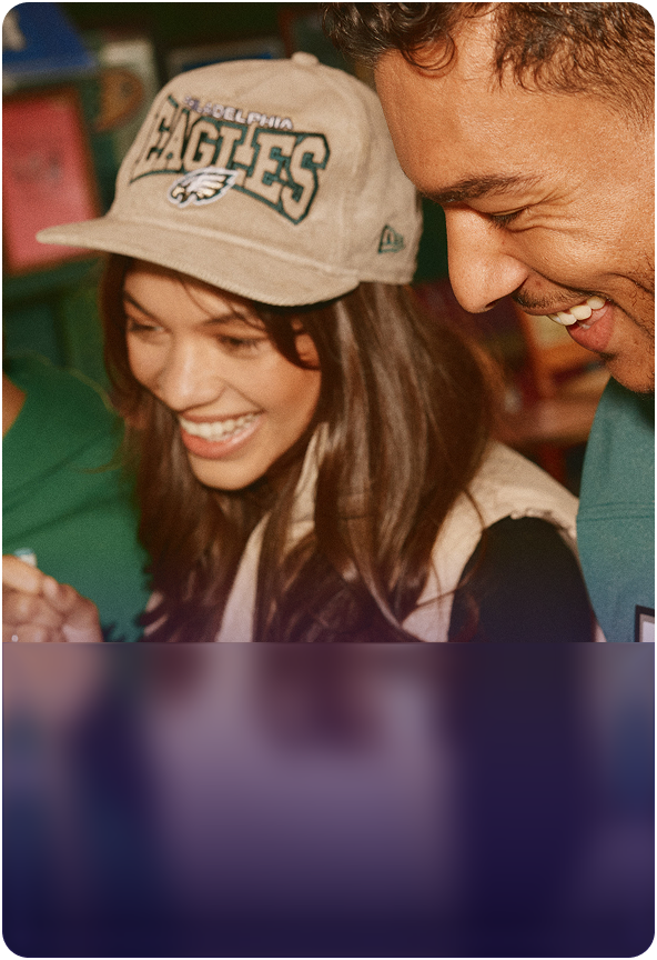 A smiling young woman wearing a tan corduroy Philadelphia Eagles New Era hat next to a laughing man while shopping for NFL apparel.