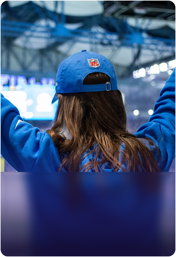 Rear view of a female fan with long brown hair wearing a blue NFL hat and matching blue hoodie cheering with her arms raised inside a football stadium during a live game.