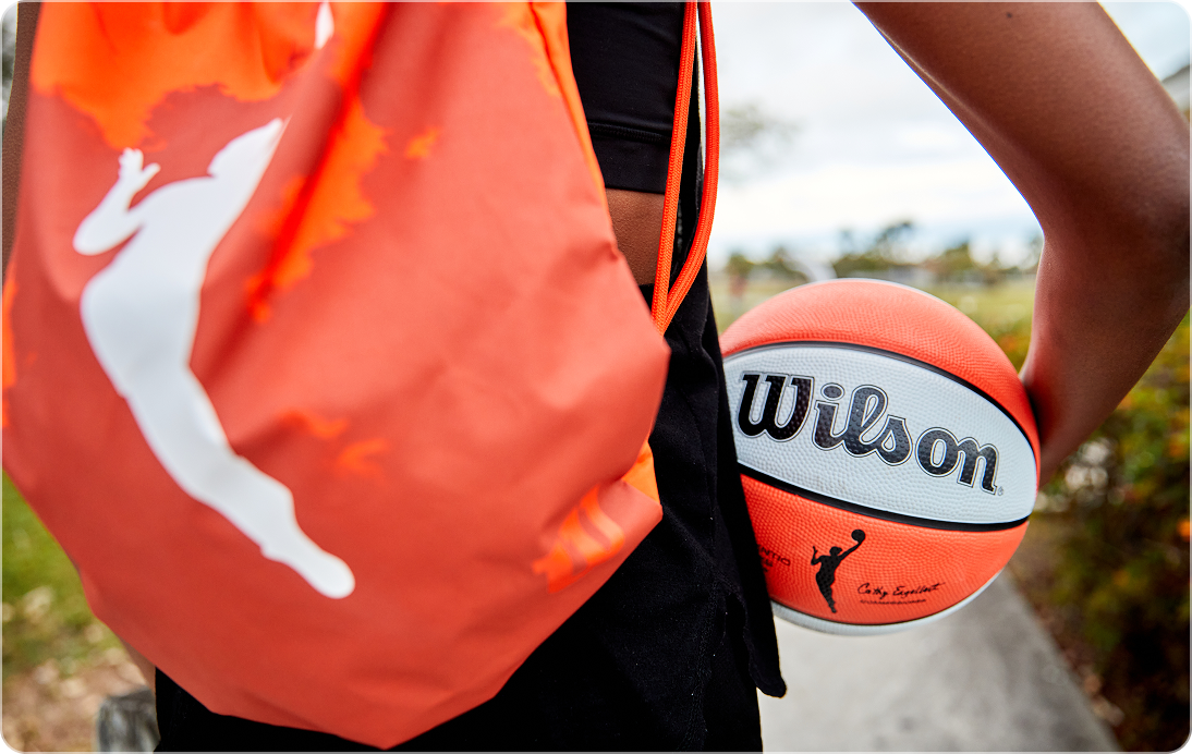 Person carrying a Wilson WNBA basketball and orange gym bag outdoors for Fanatics Sportsbook lifestyle image