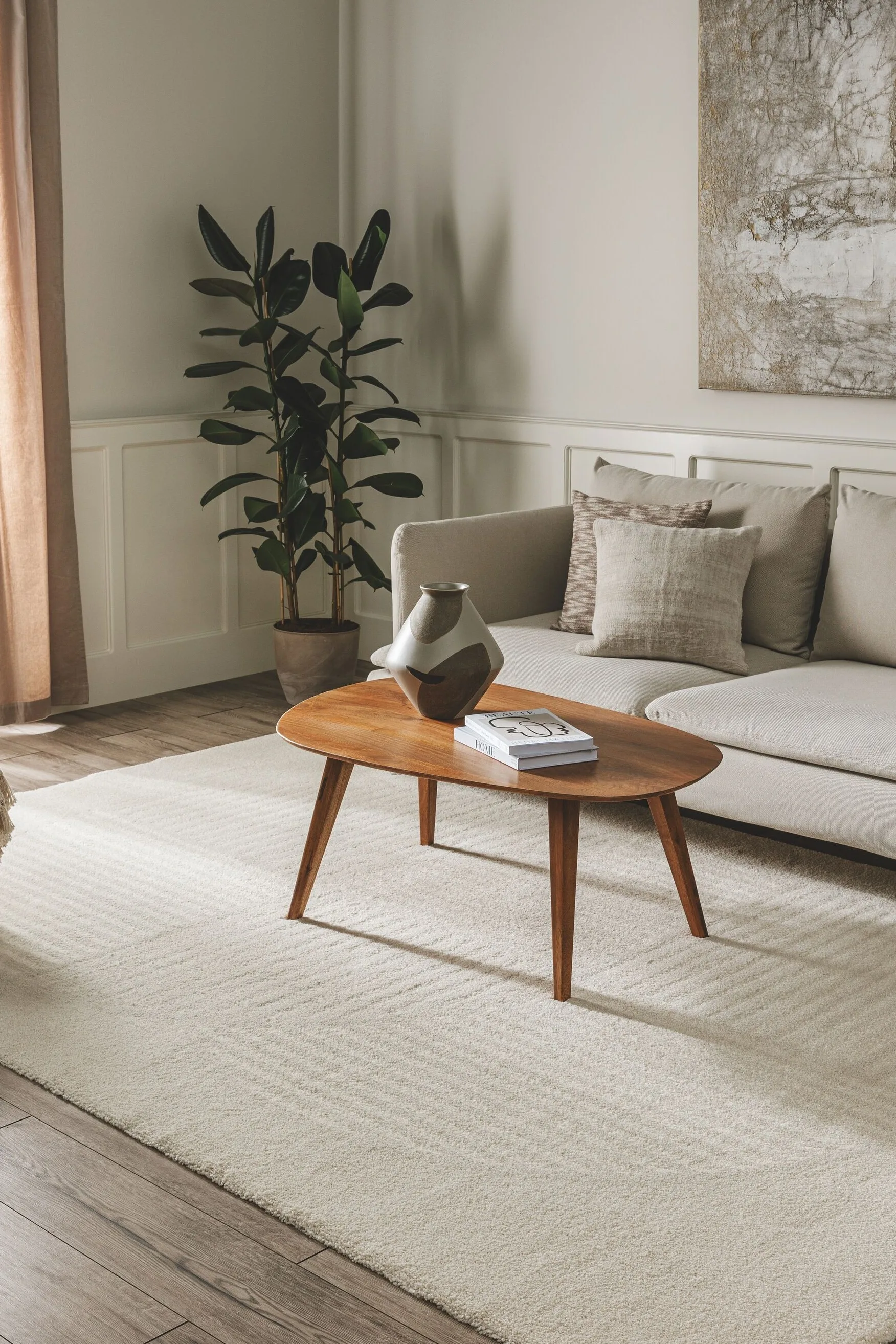 Dekoriertes Wohnzimmer in hellen Tönen mit hellgrauem Sofa und darauf abgestimmten Wohnaccessoires wie Gemälde, Vase, Pouf, Dekokissen und Teppich. Couch-Tisch aus Eiche setzt optischen Akzent als Einrichtungshighlight