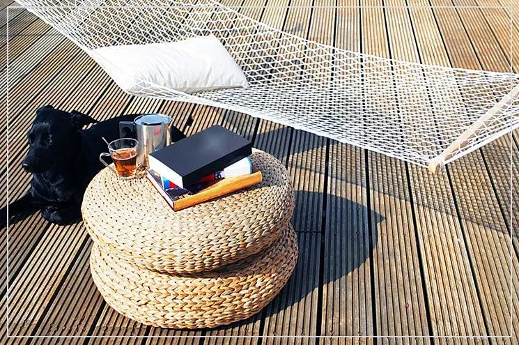 Nahaufnahm von einer Hängematte mit Kissen auf einem Balkon und einem Beistelltisch aus Jute, auf dem Getränke stehen, um entspannt Urlaub zu Hause machen zu können