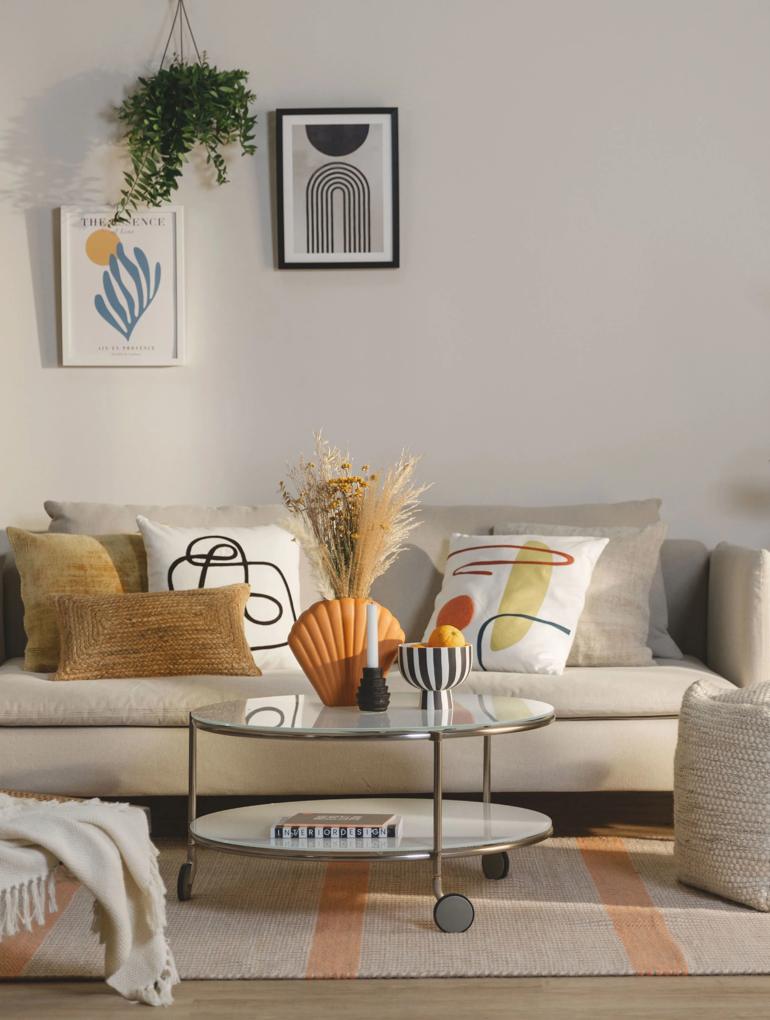 Lebhaft eingerichtetes Wohnzimmer mit verschiedenen Dekokissen in Weiß, Gelb und Braun, bunten Vasen in Orange, Bildern und einer Hängepflanze über dem Sofa