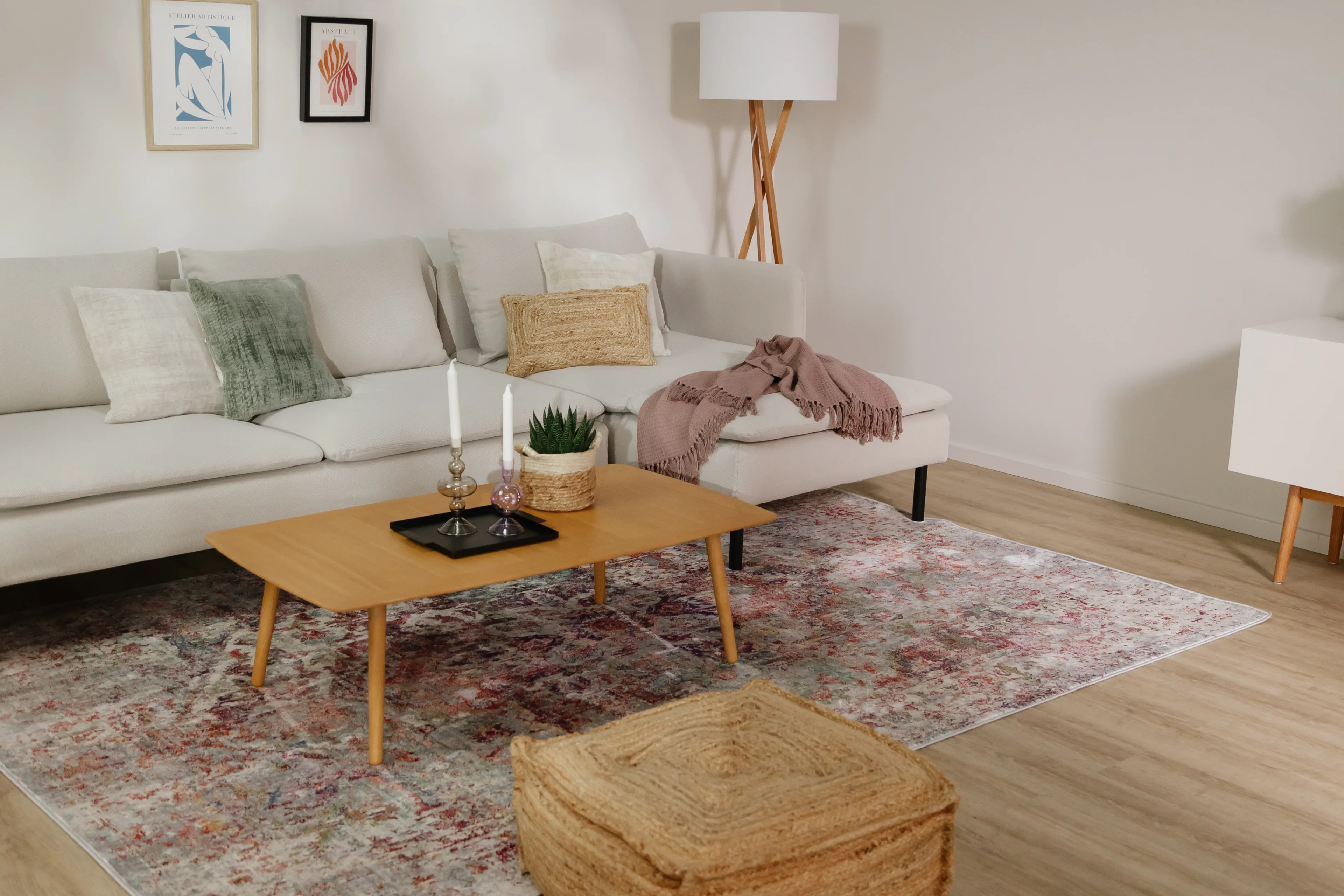 Gemütlich eingerichtetes Wohnzimmer mit hellem L-Sofa, bunten Kissen in Braun und Mintgrün, einer Kuscheldecke in Pastelllila und einem Vintage-Teppich in Pastelllila