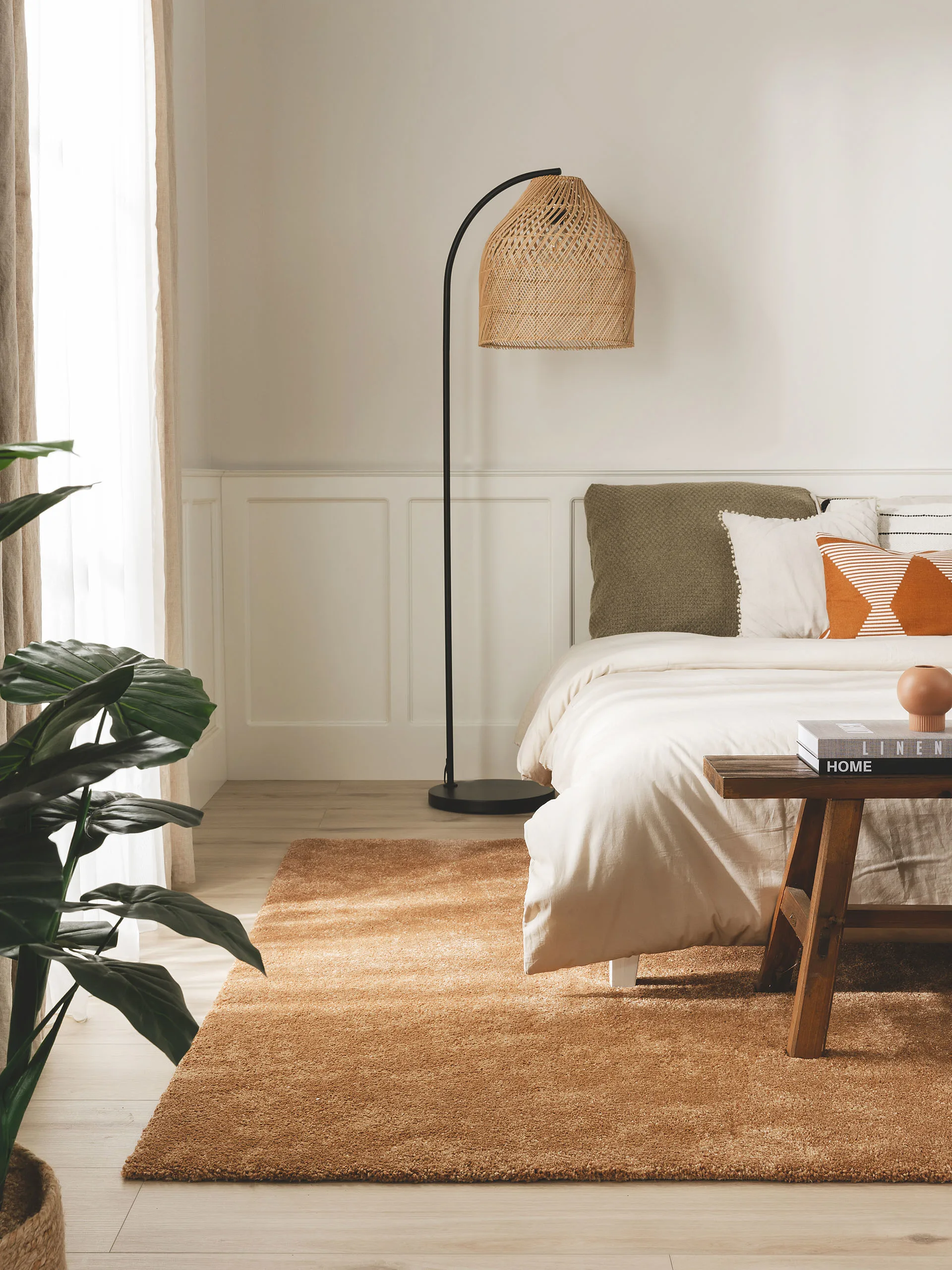 Schlafzimmer im Landhausstil mit Stehlampe aus Rattangeflecht, großem Bett dekoriert mit Kissen in Olivgrün, Orange und Weiß und einem Hochflorteppich in einem erdigen Orange-Ton
