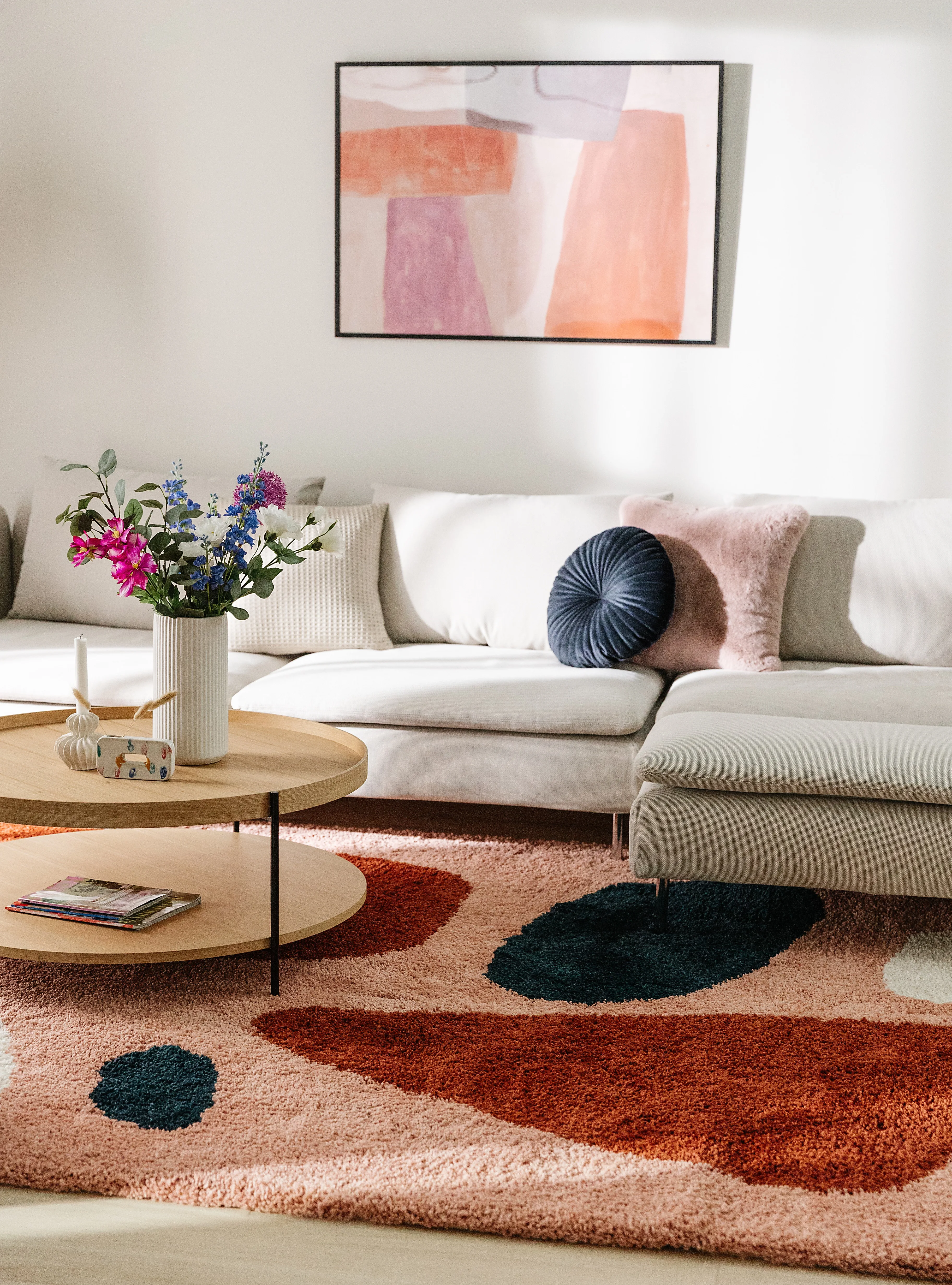 Wohnzimmereinrichtung mit farbenfrohen Dekoelementen wie Kissen, Teppich und Bild in Rot, Rosa, Lila und Orange und einer schlichten Couch in Creme