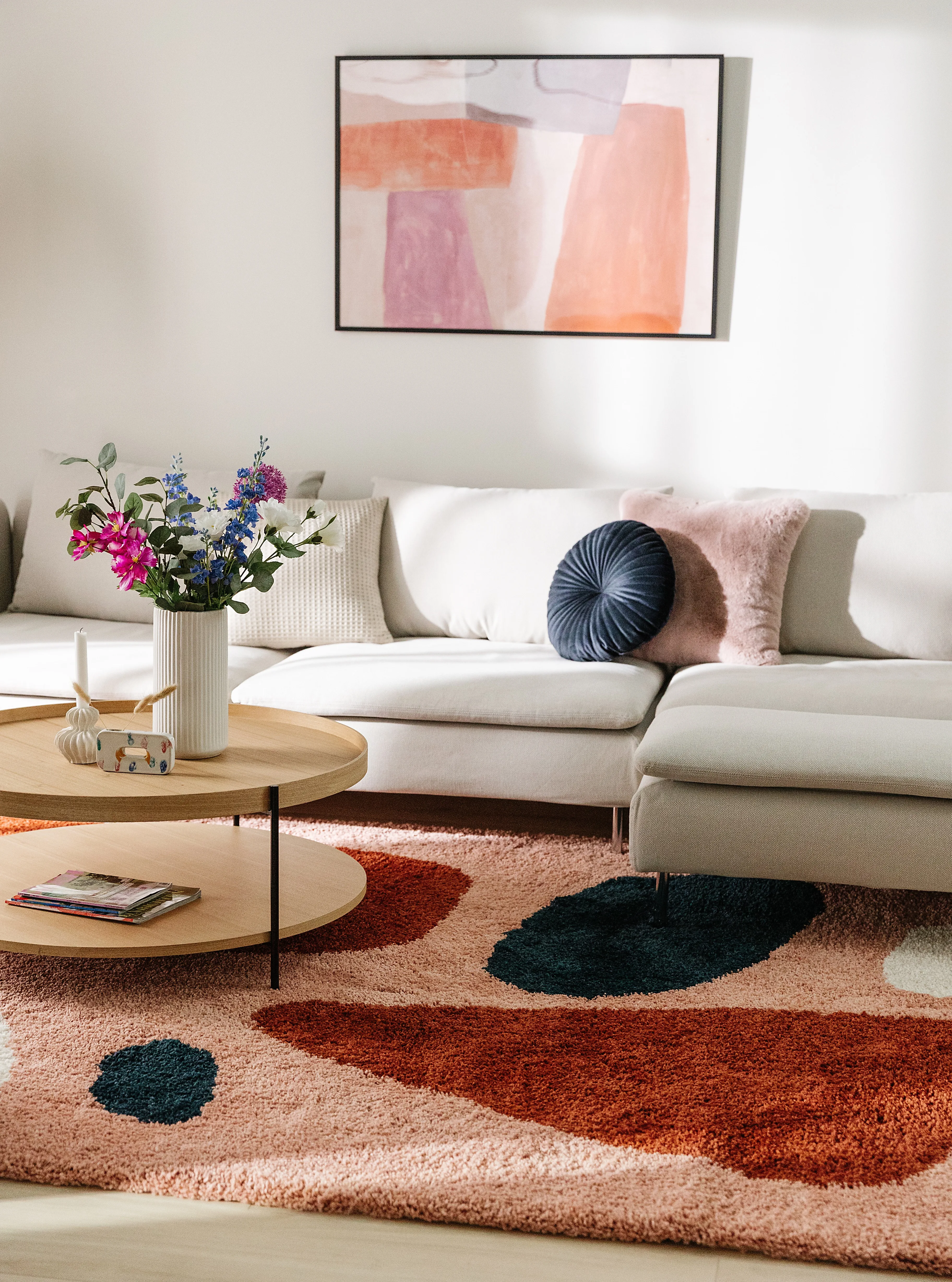 Wohnzimmereinrichtung mit farbenfrohen Dekoelementen wie Kissen, Teppich und Bild in Rot, Rosa, Lila und Orange und einer schlichten Couch in Creme