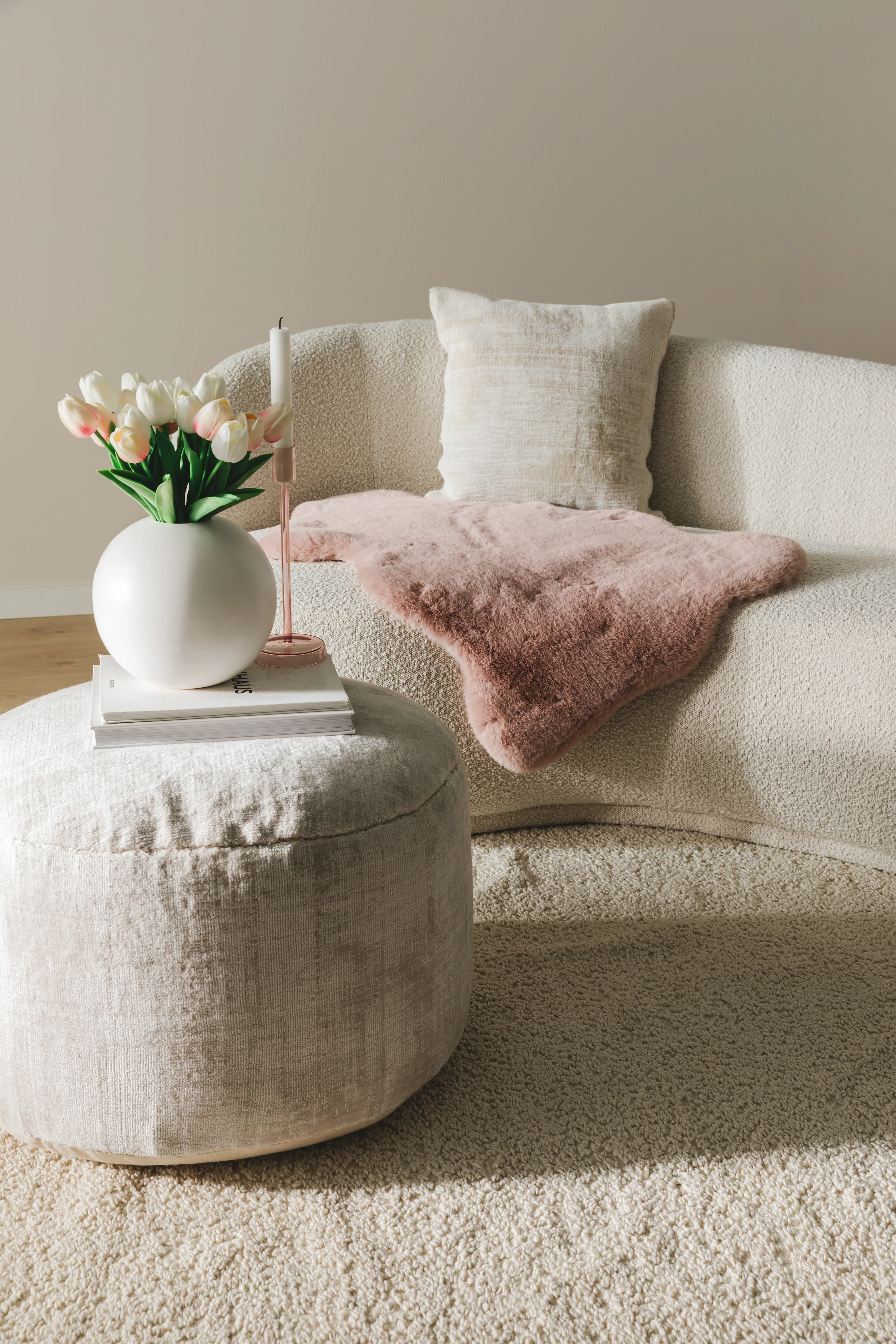 Weiches Deko-Kunstfell in Rosa drapiert auf einer cremefarbenen Couch in S-Form hinter einem hellen Pouf, auf dem Bücher und eine helle Vase mit weißen Tuplen als Valentinstagsgeschenk stehen