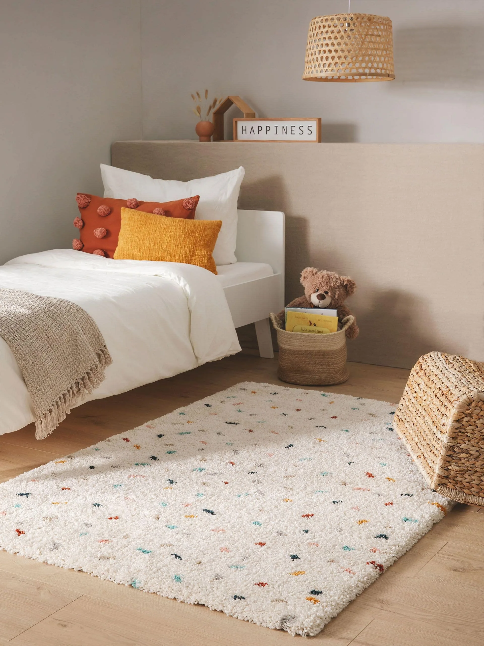 Kinderzimmereinrichtung mit einem weißen Teppich mit bunten Punkten, farbenfrohe Kissen auf einem Einzelbett und einer Spielzeugtruhe und einem Hanf-Korb in der Ecke, in dem ein Kuscheltier und Bücher verstaut wurden