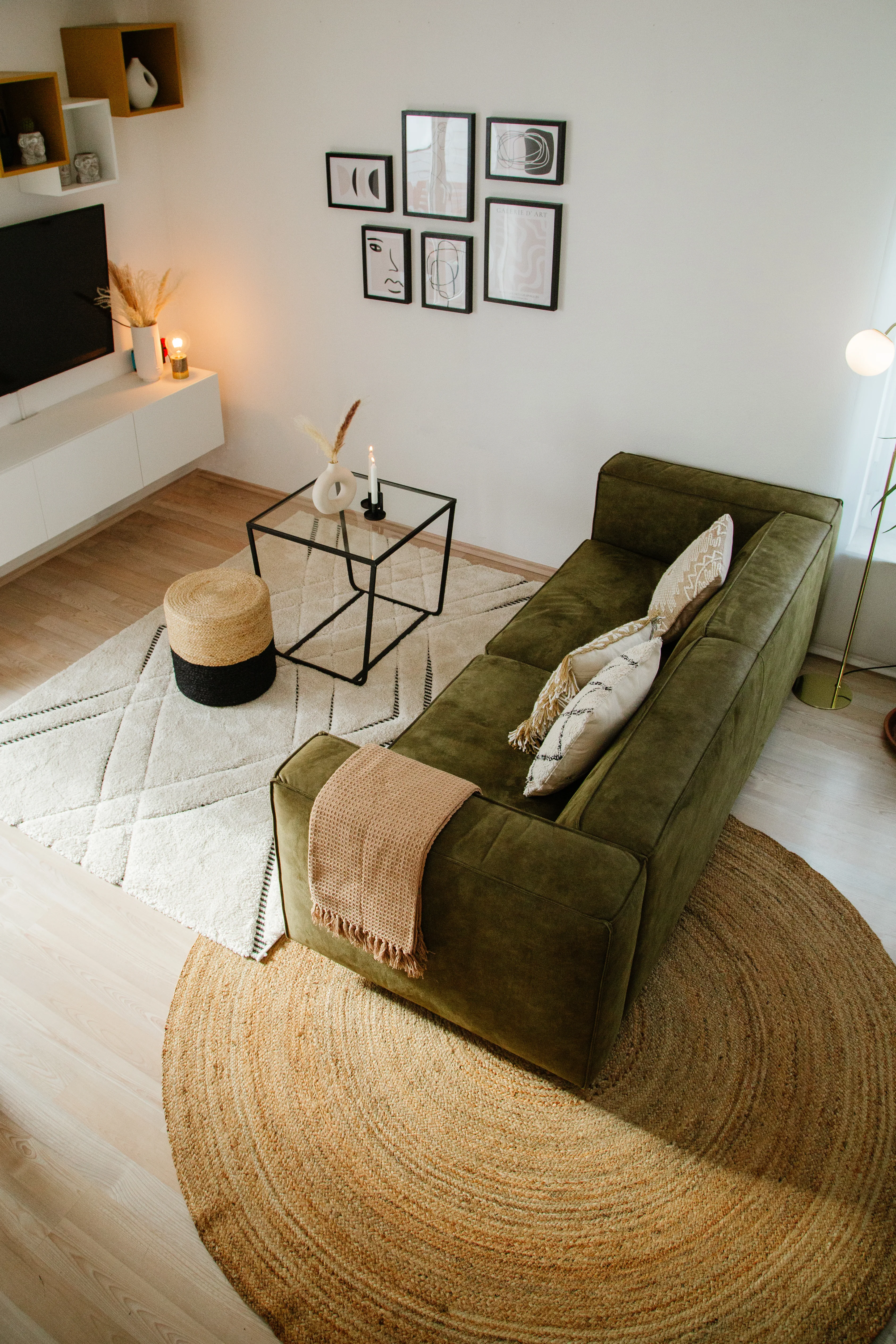 Voll eingerichtetes Wohnzimmer von oben mit olivgrüner Couch, einem weißen und einem braunen Teppich übereinander gelayert und einer weißen TV-Kommode