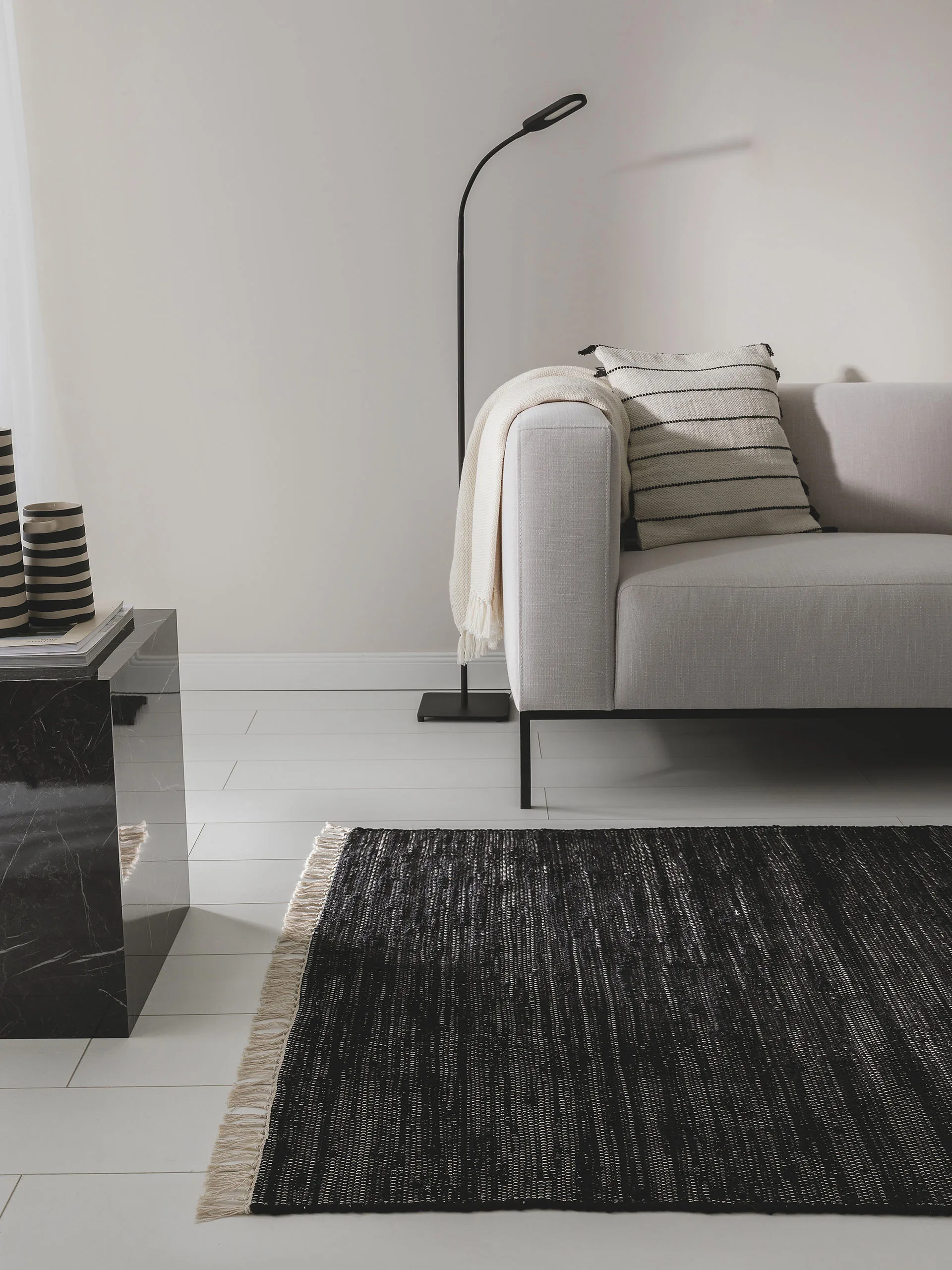 Dunkles, luxuriöses Wohnzimmer mit grauem Sofa, schwarzem Beistelltisch aus Marmor und schwarzem Teppich mit Fransen auf weißen Fliesen
