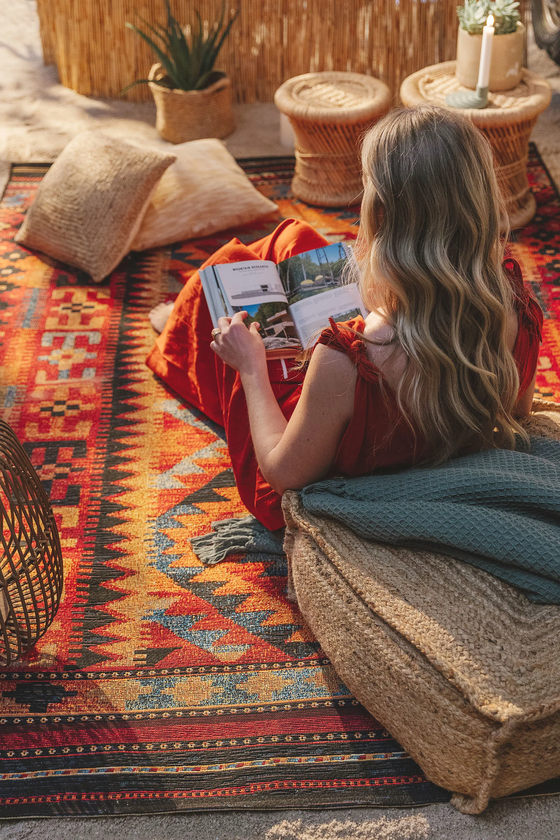 Blonde Frau, die draußen vor ihrem Van auf einem Kelim-Teppich und angelehnt an einen Jute-Pouf ein Buch in der Sonne liest
