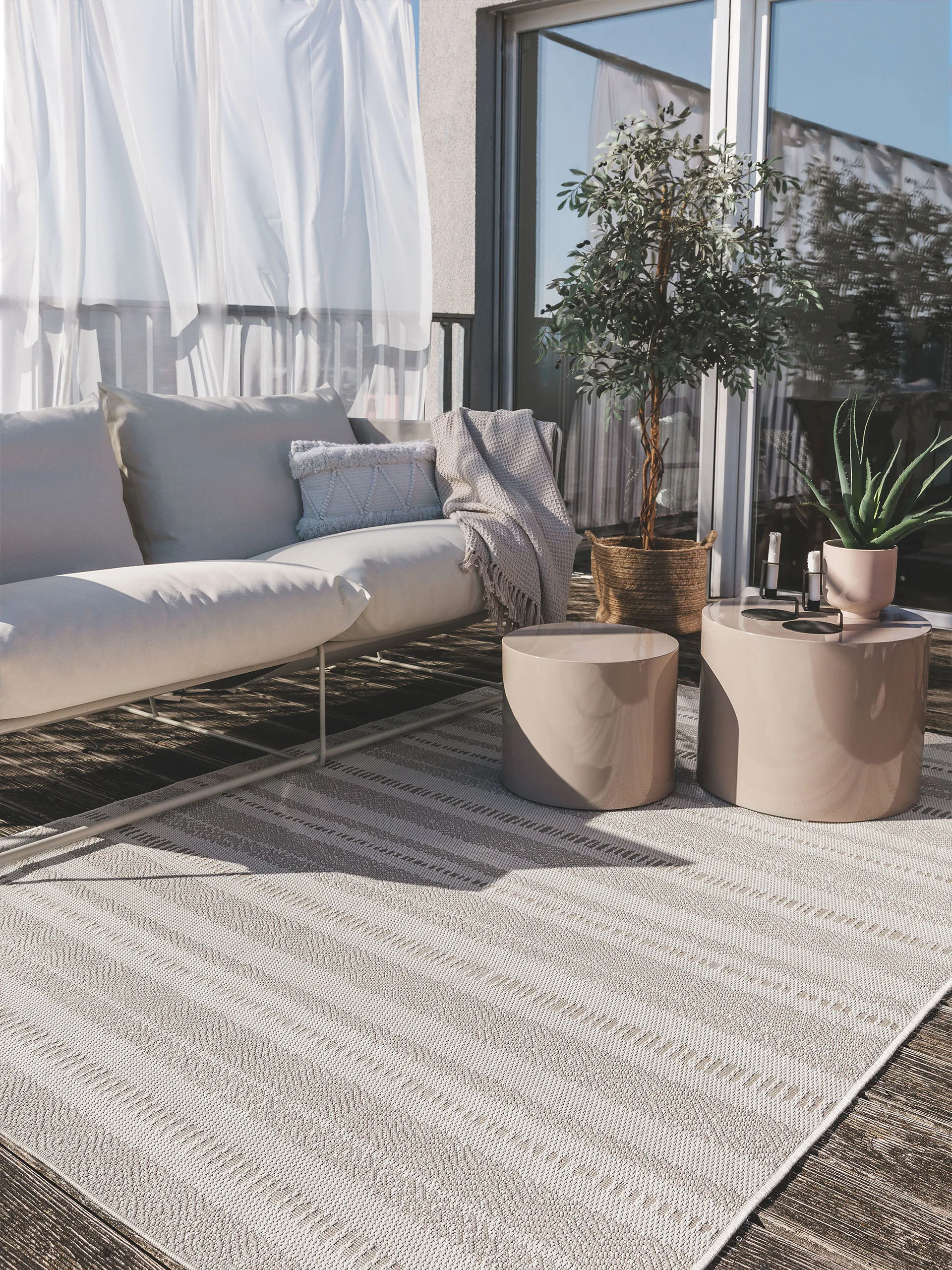 Helle Gartendeko mit grauem, großen Outdoor-Teppich, grauem Sofa und zwei beigen Beistelltischen und einer großen Topfpflanze mit weißen Blüten