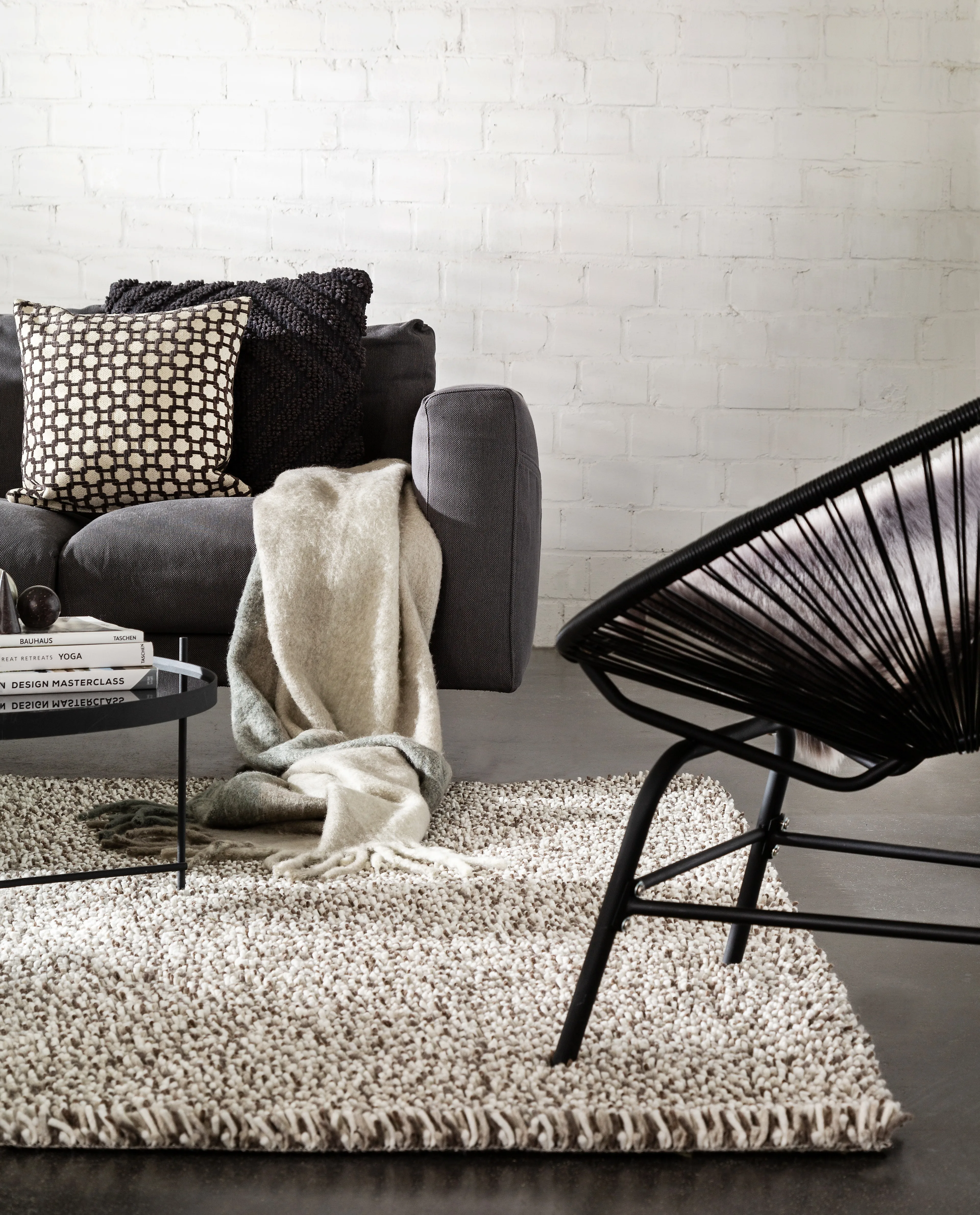 Wohnzimmer im schicken Industrial Style mit schwarzem Sofa, schwarzem Sessel, schwarzem Couch-Tisch und schwarz-weißen Dekoelementen wie drei Kissen, ein Hochflorteppich und einer weichen Decke