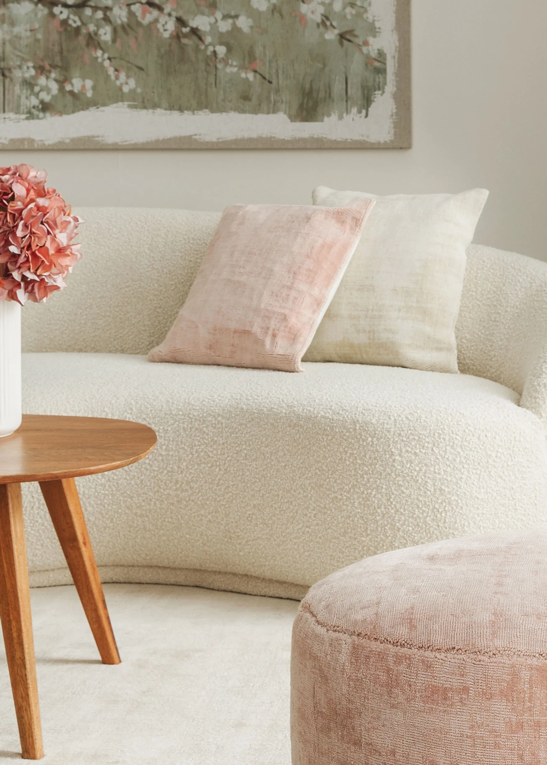 Zwei Dekokissen in Weiß und Rosa auf einem cremefarbenen Sofa in organischer Form, vor dem ein rosa Pouf und ein brauner Couchtisch mit rosanen Blumen in einer weißer Vase steht