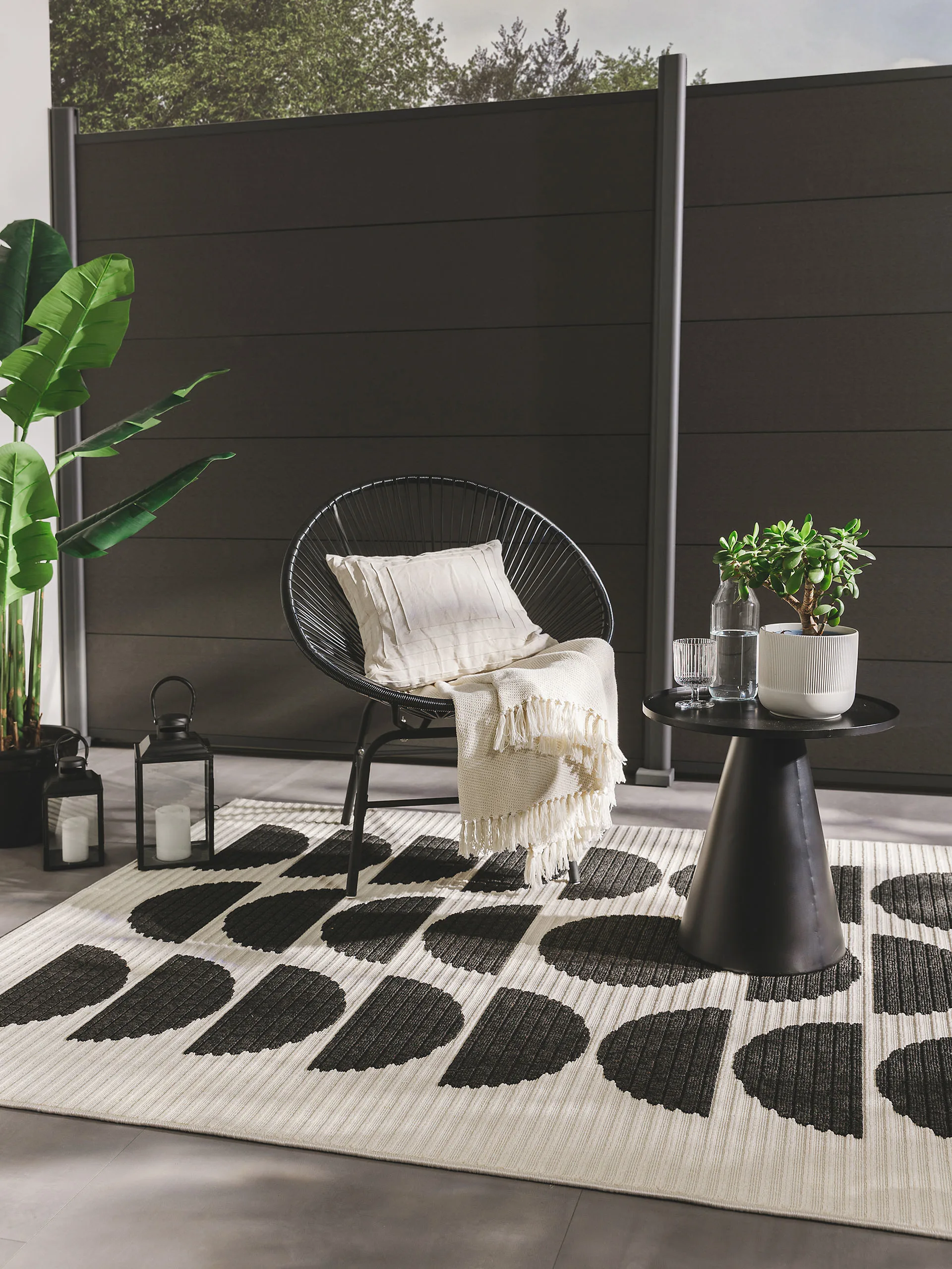 Stillvolle Gartendeko mit schwarz-weißem Outdoor-Teppich und einem schwarzen Sessel mit Beistelltisch und günstigen Dekoelementen wie Laternen, Kissen, Decken und Pflanzen