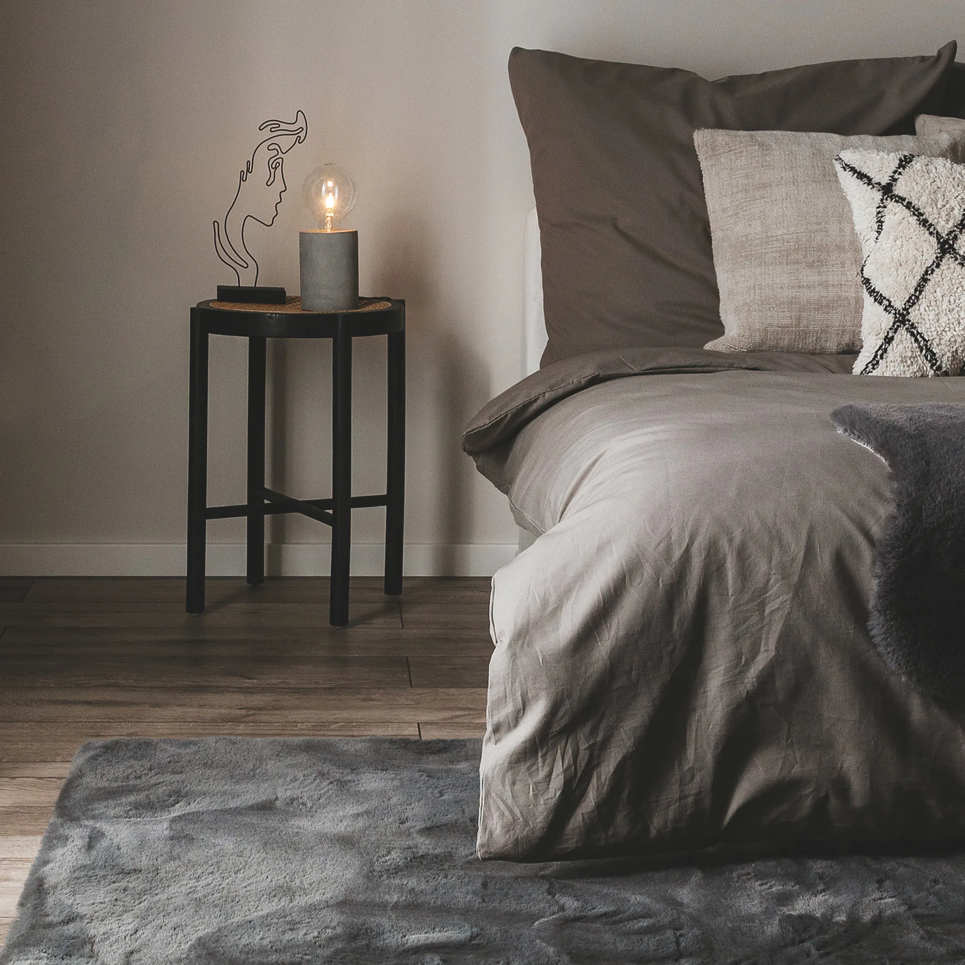 Schlafzimmer im Industrial Style mit grauer Bettwäsche, einem weichen Shaggy-Teppich in Grau als Bettvorleger und einem schwarzen Nachtschrank, auf dem eine Stehlampe im rustikalen Glühbirnen-Design steht