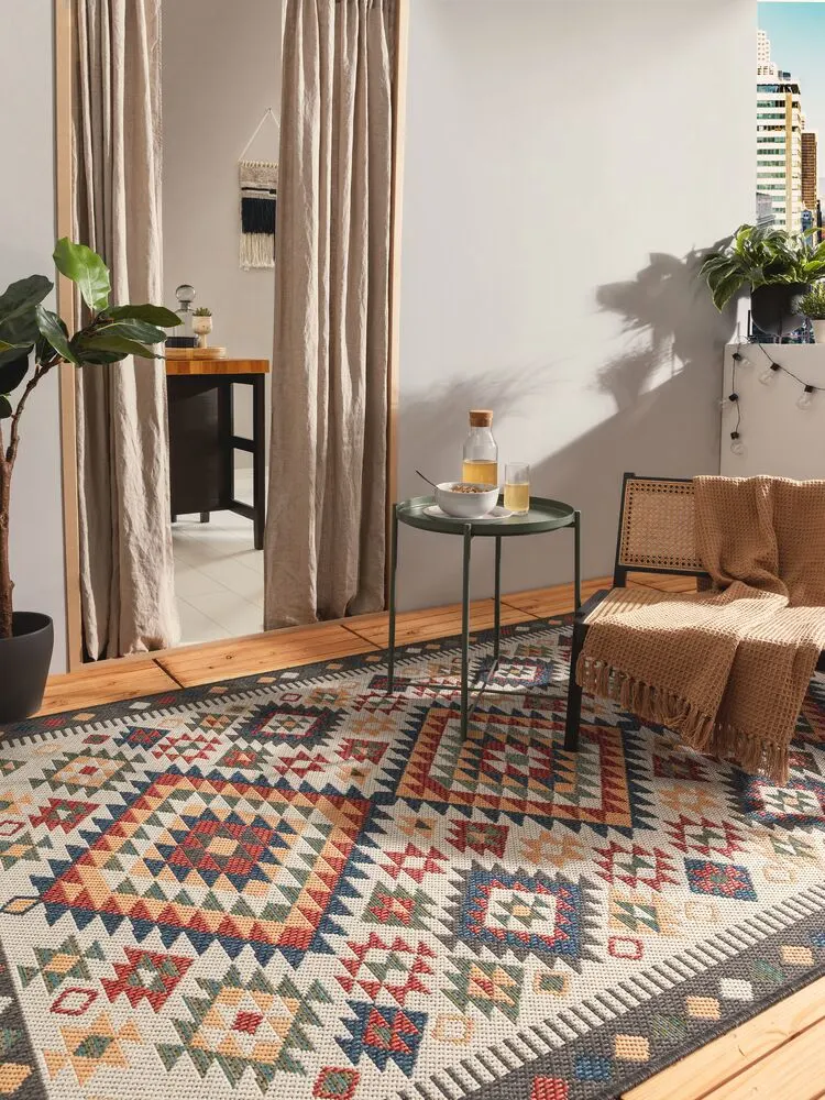 Balkon dekoriert für den Frühling mit buntem Outdoor-Teppich, Boho-Pouf, gemütlicher Hängematte mit Bommeln und grünen Pflanzen