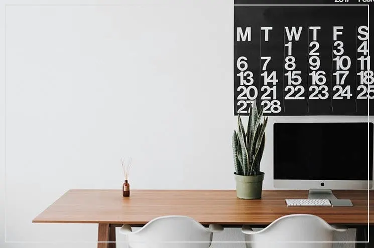 Büro eingerichtet mit einem aufgeräumten Schreibtisch aus Holz mit Desktop, Tastatur und Pflanze, zwei Schreibtischstühlen in Weiß und einem großen, schwarzen Terminkalender an der Wand