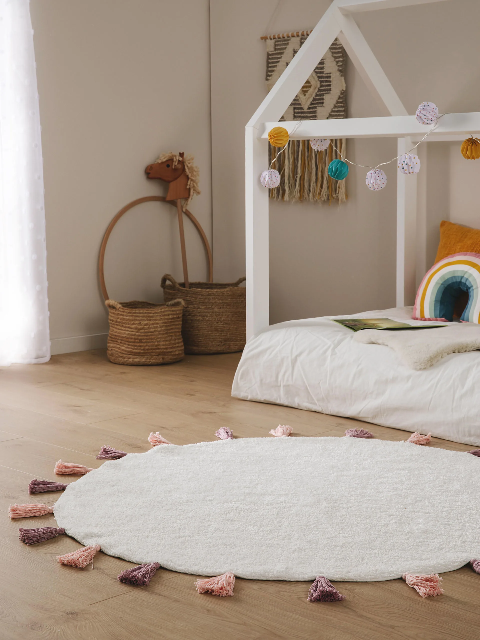 Kinderzimmer gestaltet mit einem runden, weißen Teppich mit Bommeln, einem Himmelbett und verspielter Dekoration wie einer bunten Lichterkette über und ein Regenbogenkissen auf dem Bett