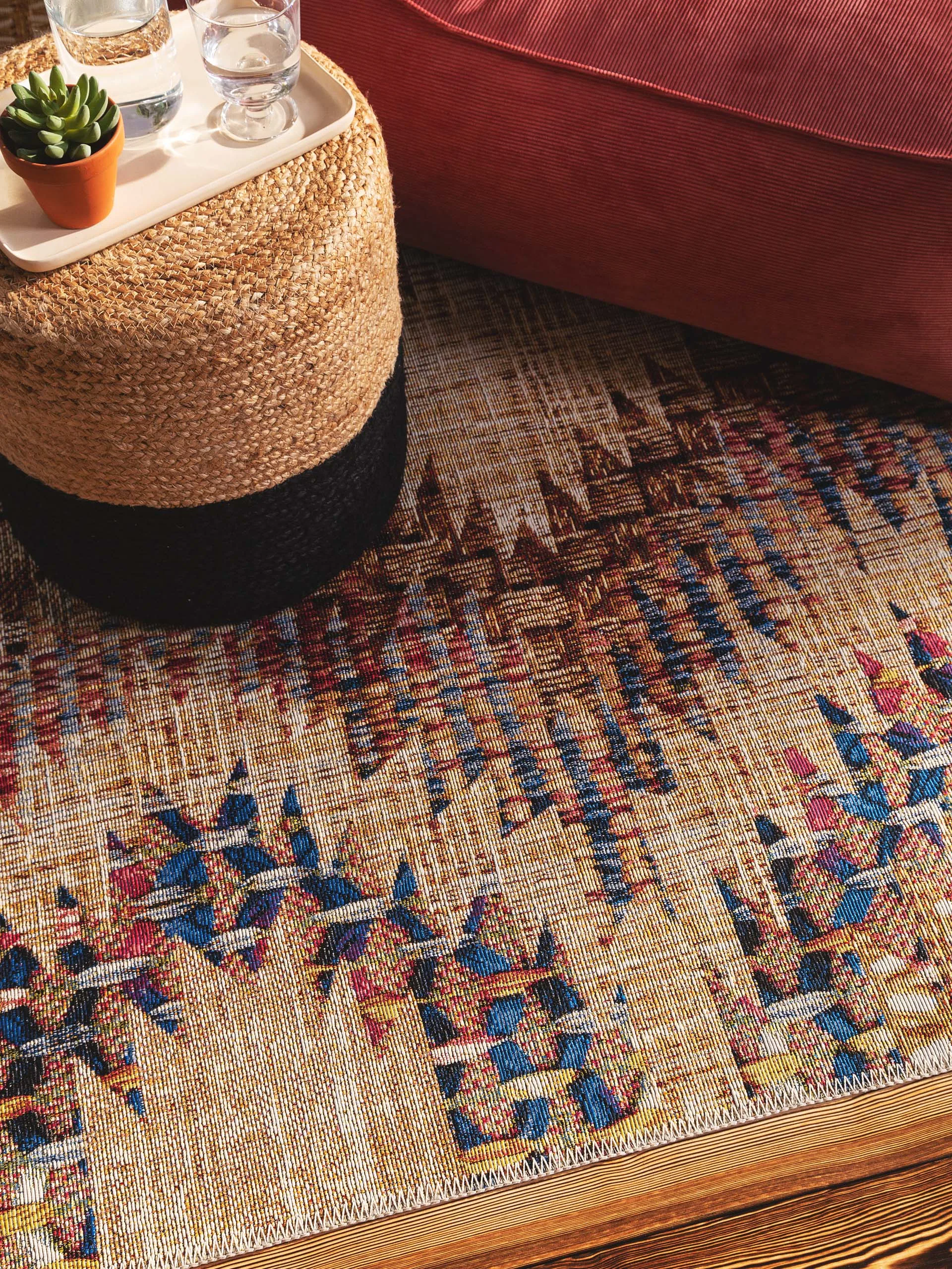 Detailaufnahme von einem Outdoor-Teppich im bunten Kelim-Design, auf dem ein Pouf aus Jute mit Tablett und einem Glas Wasser steht
