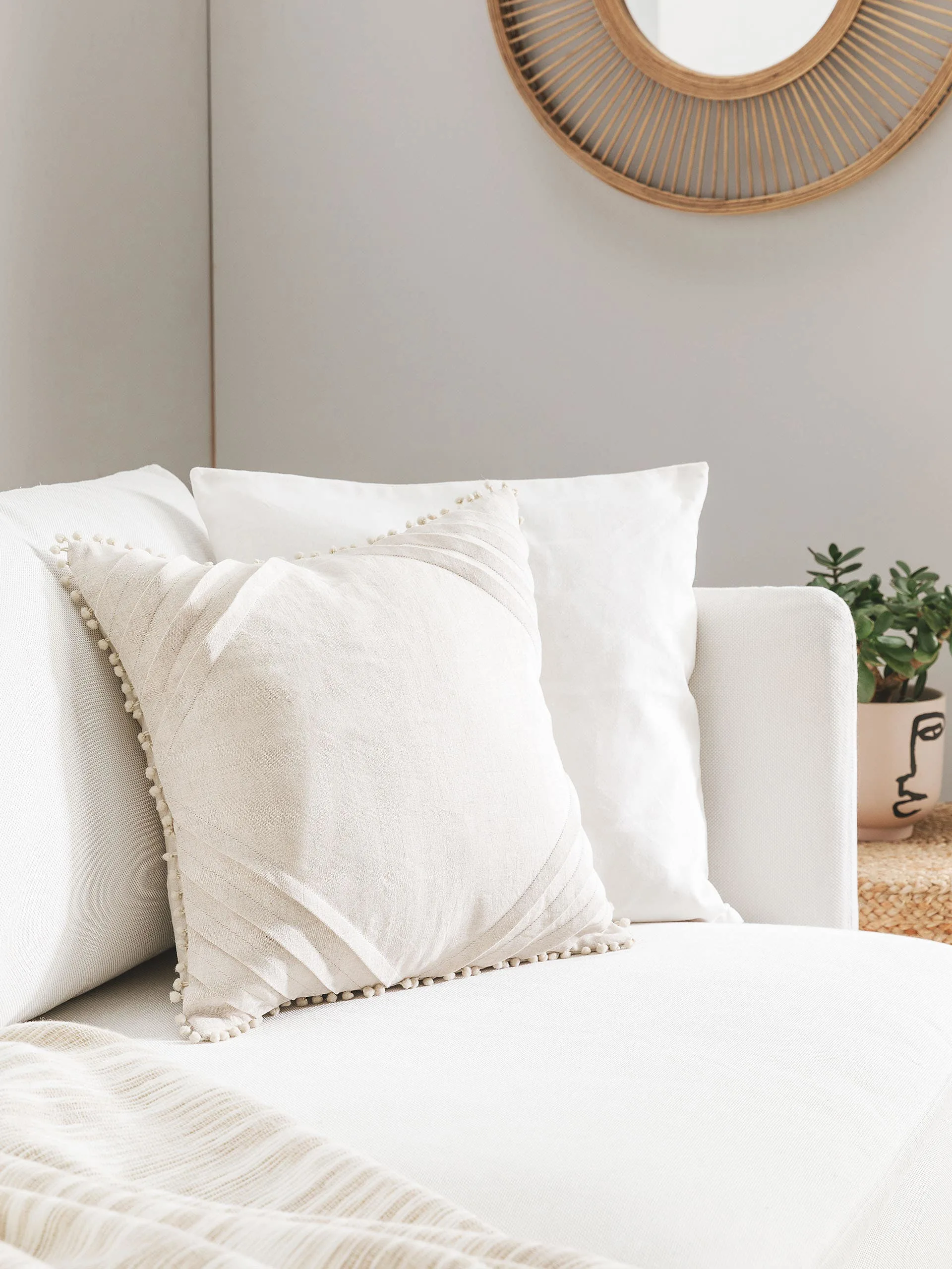 Detailaufnahme von zwei weißen Dekokissen mit Bommeln auf einem weißen Sofa in einem kleinen Wohnzimmer mit hellen Wänden