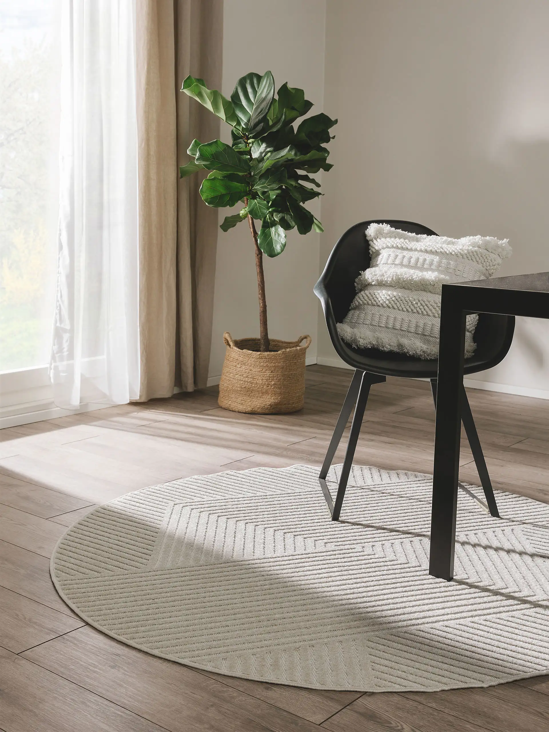 Kleiner, runder Teppich unter einem schwarzen Sessel mit weißem Boho-Kissen und einer große Pflanze in der linken Zimmerecke