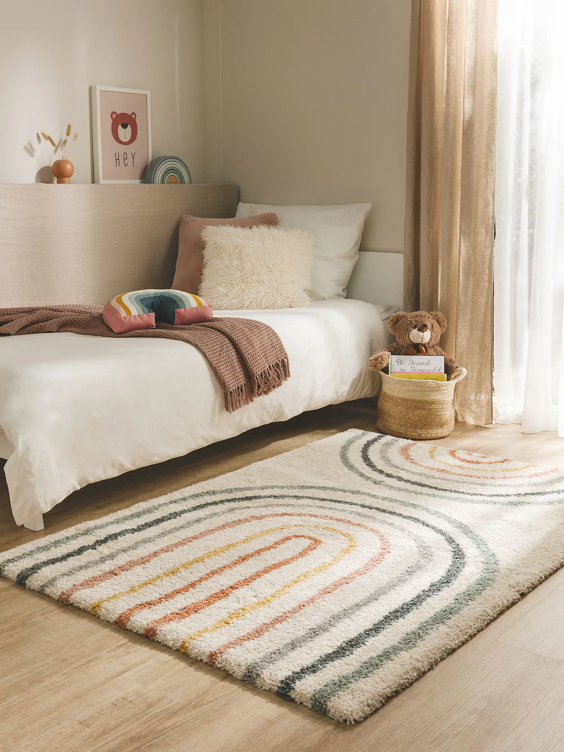 Kleiner, bunter Teppich im Kinderzimmer mit Regenbogen-Design vor einem Kinderbett, auf dem Kissen, eine Kuscheldecke und ein Kuscheltier liegen