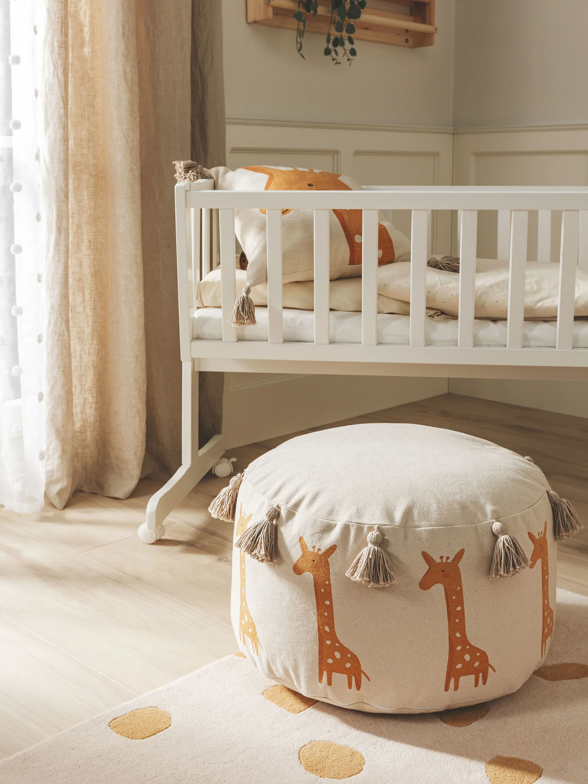 Kinderzimmer eingerichtet im Safari-Look mit einem Pouf im Giraffen-Design vor einem Baby-Gitterbett und einem Giraffen-Kissen im sowie einem beigen Teppich mit gelben Tupfern vor dem Bett