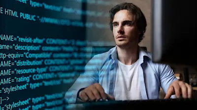 A man sits at a computer, focused on a screen displaying lines of code.