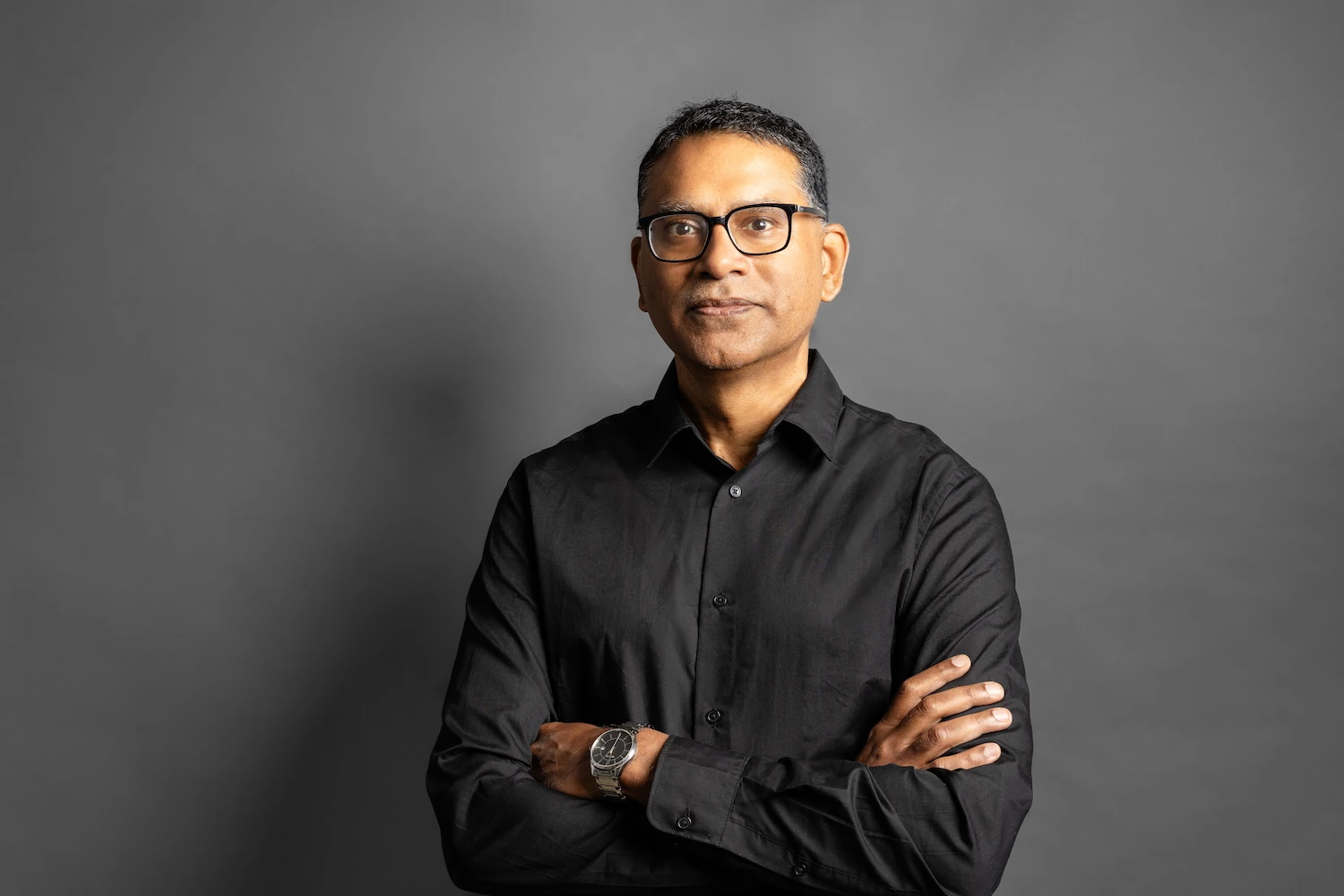 Professional portrait of a person in a black button-up shirt with arms crossed against a gray background.