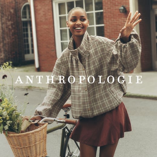 Model in a plaid bomber jacket and brown pleated skirt waving next to a bicycle with a flower-filled basket.