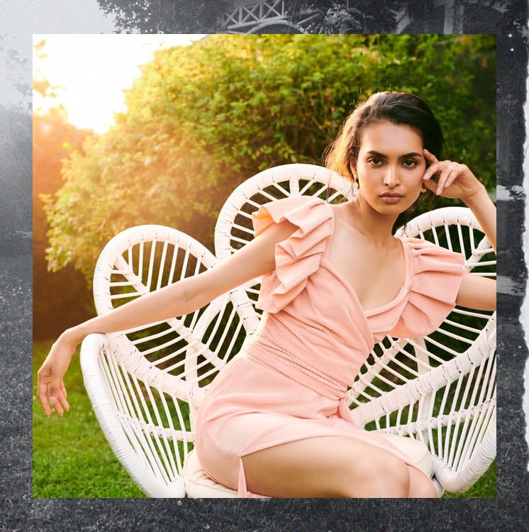 Model in pink, pleated party dress sitting in a white chair.