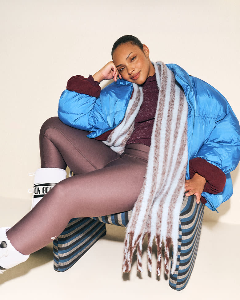 Model wearing a bright blue puffer, leggings, fringed scarf, and snow boots, lounging in a striped chair.