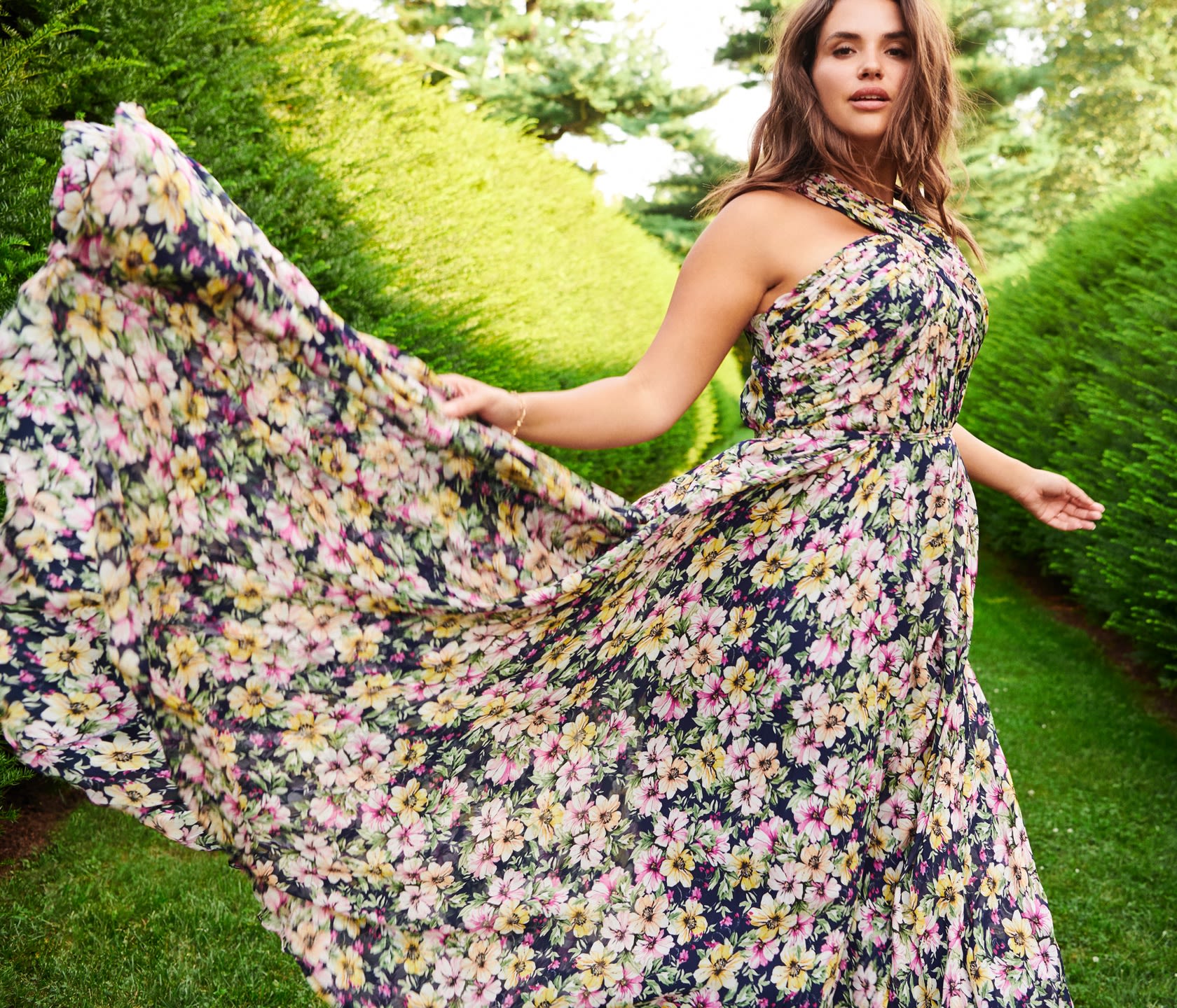 Model with a floral, colorful, and windswept dress walking through a graden.