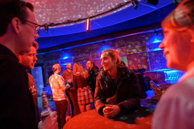 A group of two women and two men stand around a bar table and talk. The room is lit with colored lights.