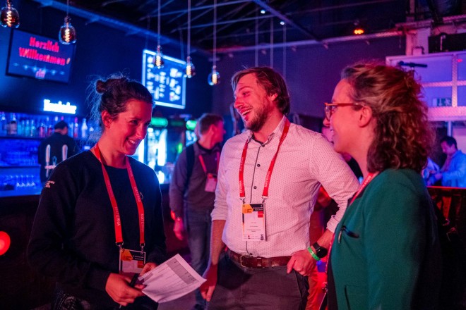 A group of two women and two men stand around a bar table and talk. The room is lit with colored lights.