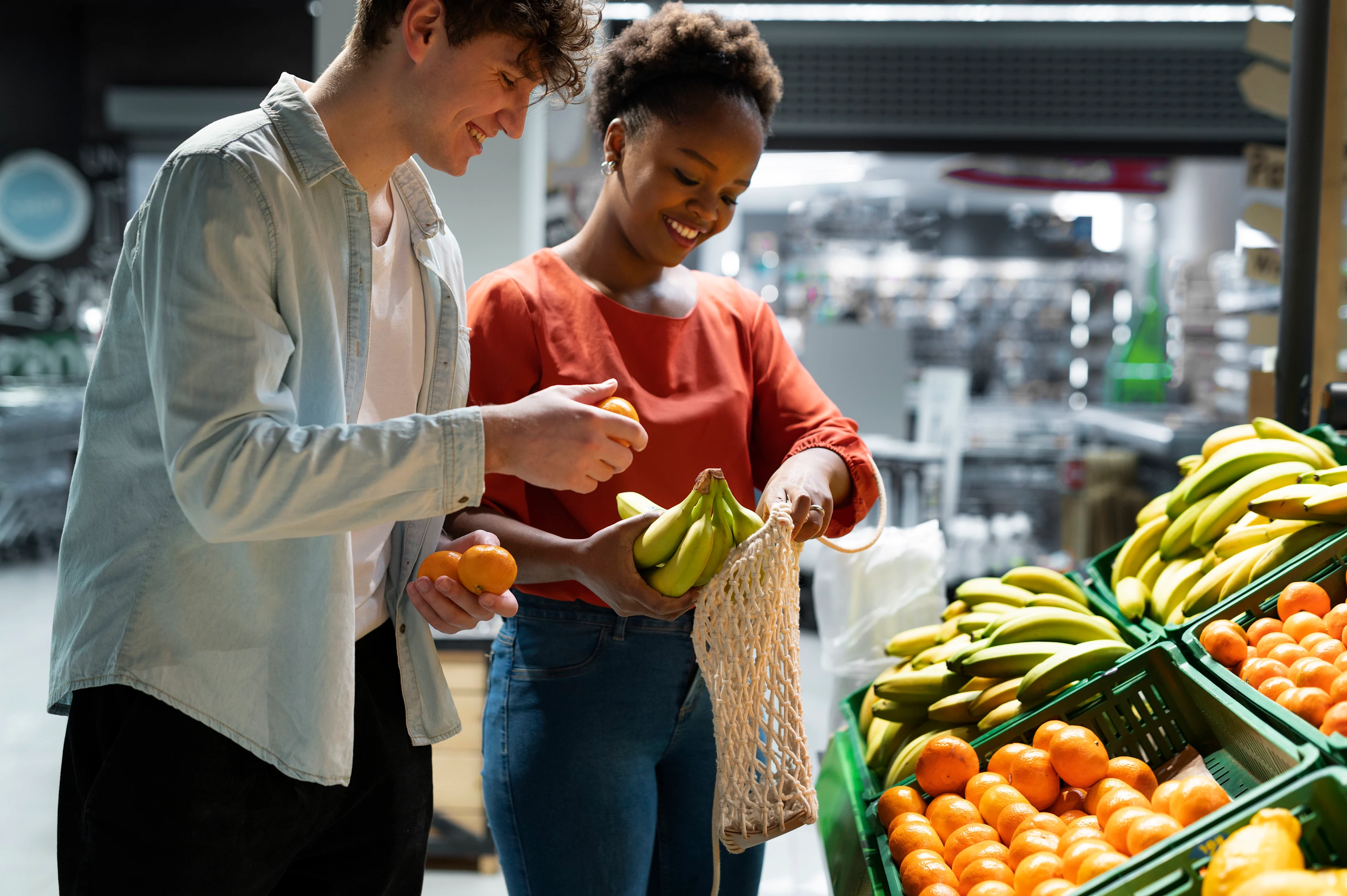 Dos personas sonríen mientras colocan plátanos y naranjas en una bolsa de malla frente a otras frutas y verduras.