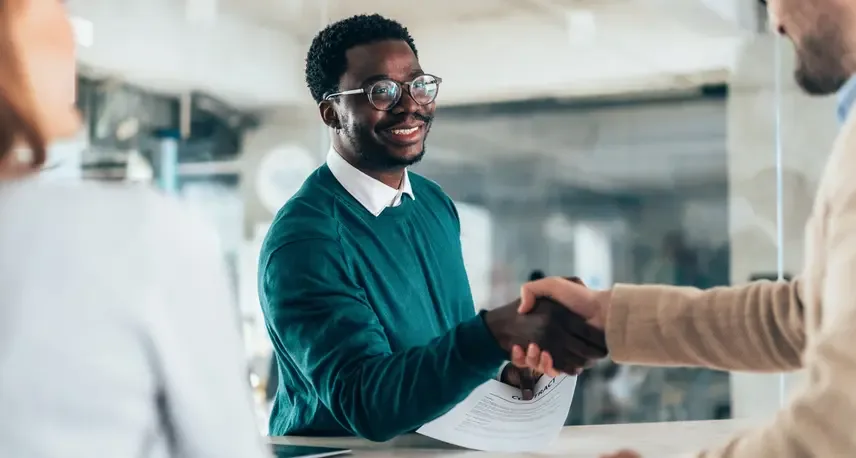 A person with dark hair and a teal shirt shaking the hand of another person while smiling