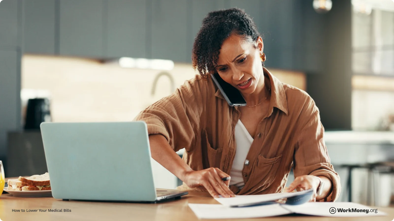 A woman looks stressed while talking on the phone, reviewing documents at a kitchen counter. She is multitasking with a laptop open in front of her, a pen in hand, and a sandwich and orange juice nearby. The scene reflects the challenge of managing medical bills.